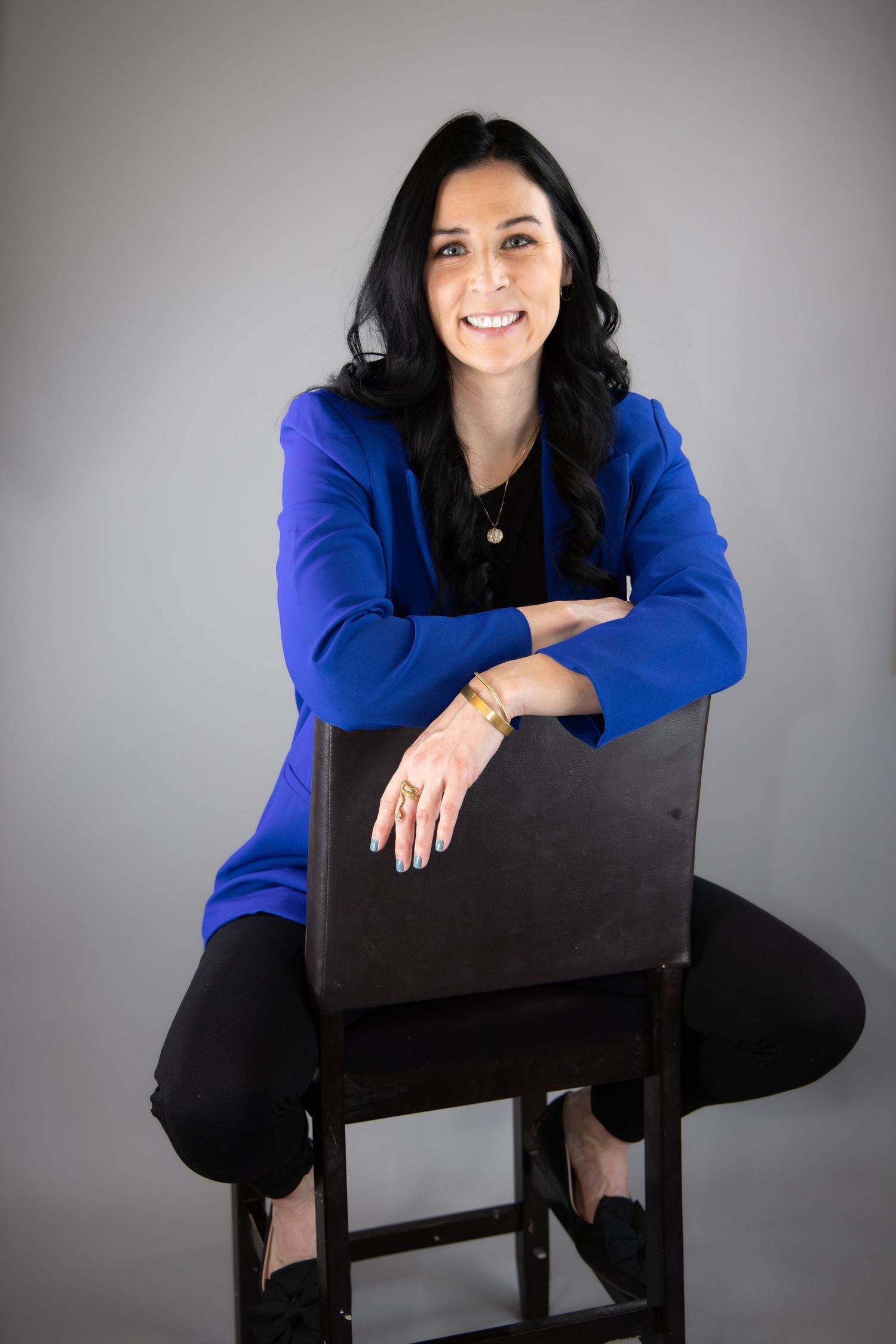 Woman in blue blazer and black pants sits confidently on a dark wooden chair against a gray background.