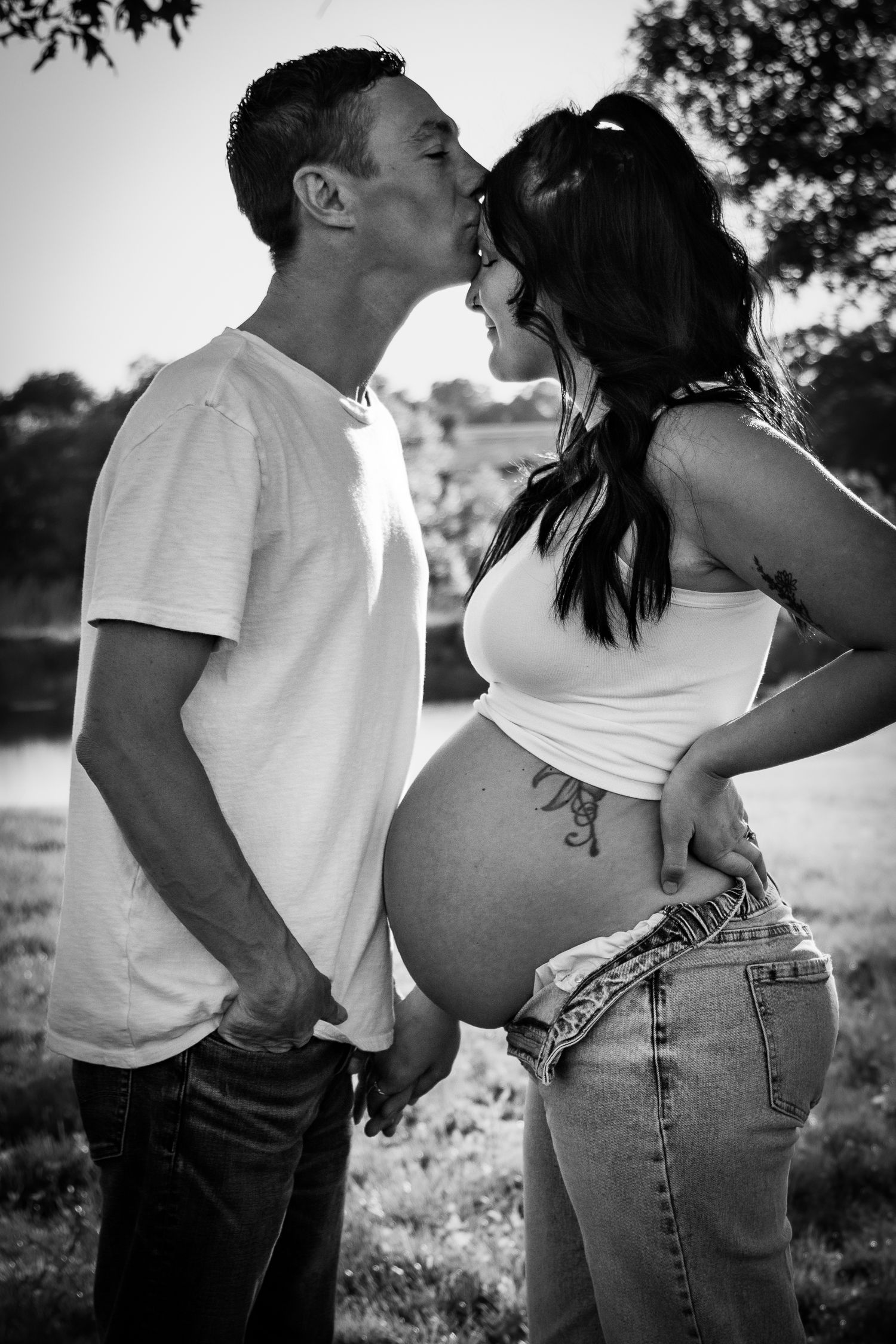 A black and white photo of a man kissing a pregnant woman outdoors near a pond.