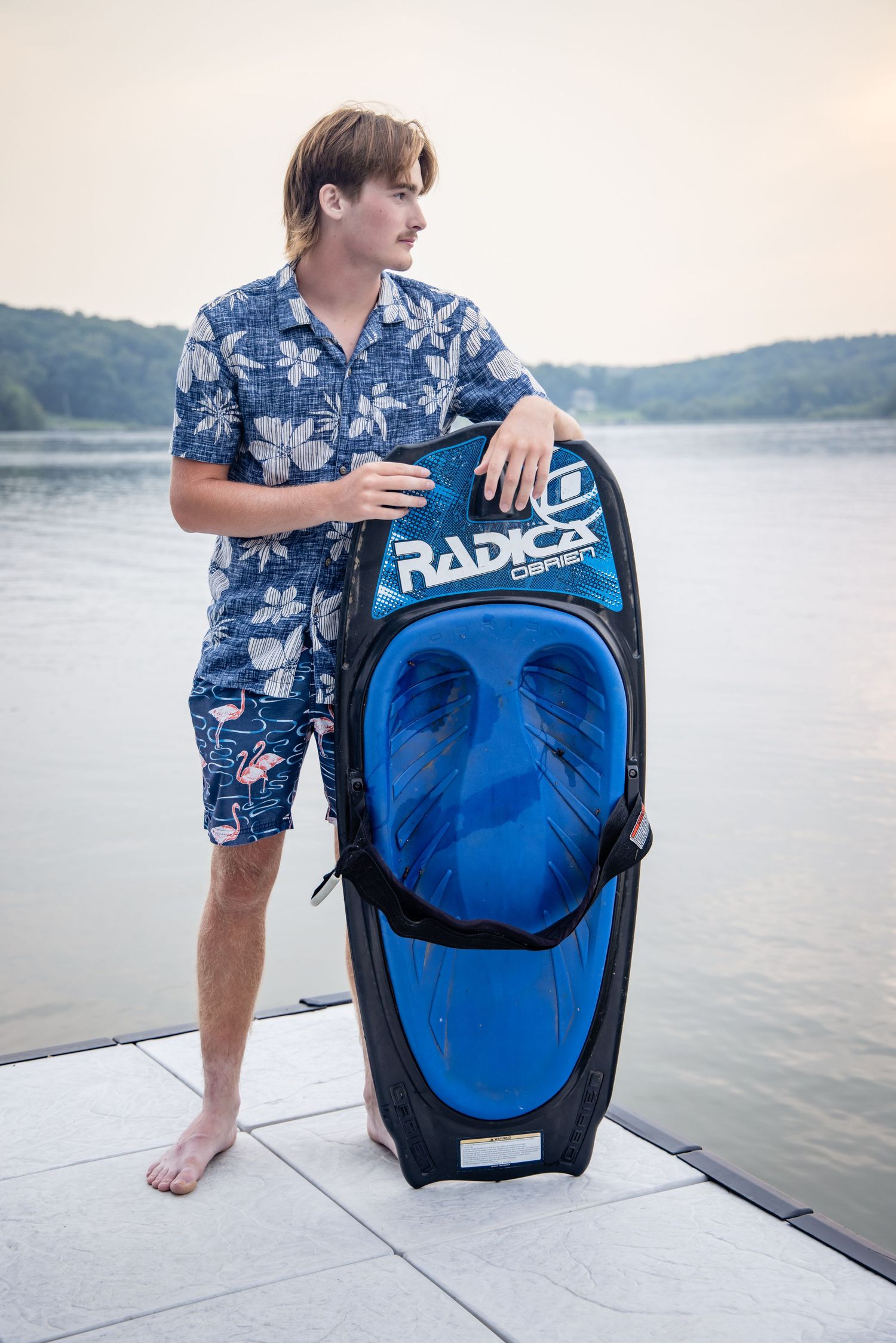 Young man in floral shirt holding a Radical Connelly kneeboard bag on a dock by a lake.