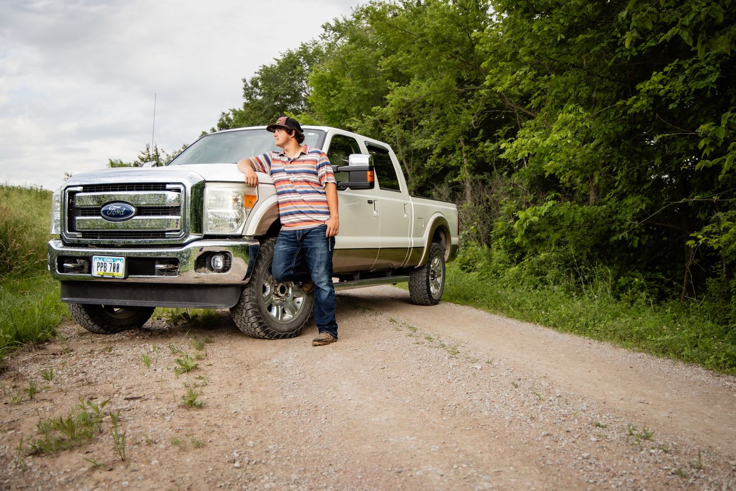 Man leaning against a Ford F-250 truck on a rural gravel road surrounded by green trees.