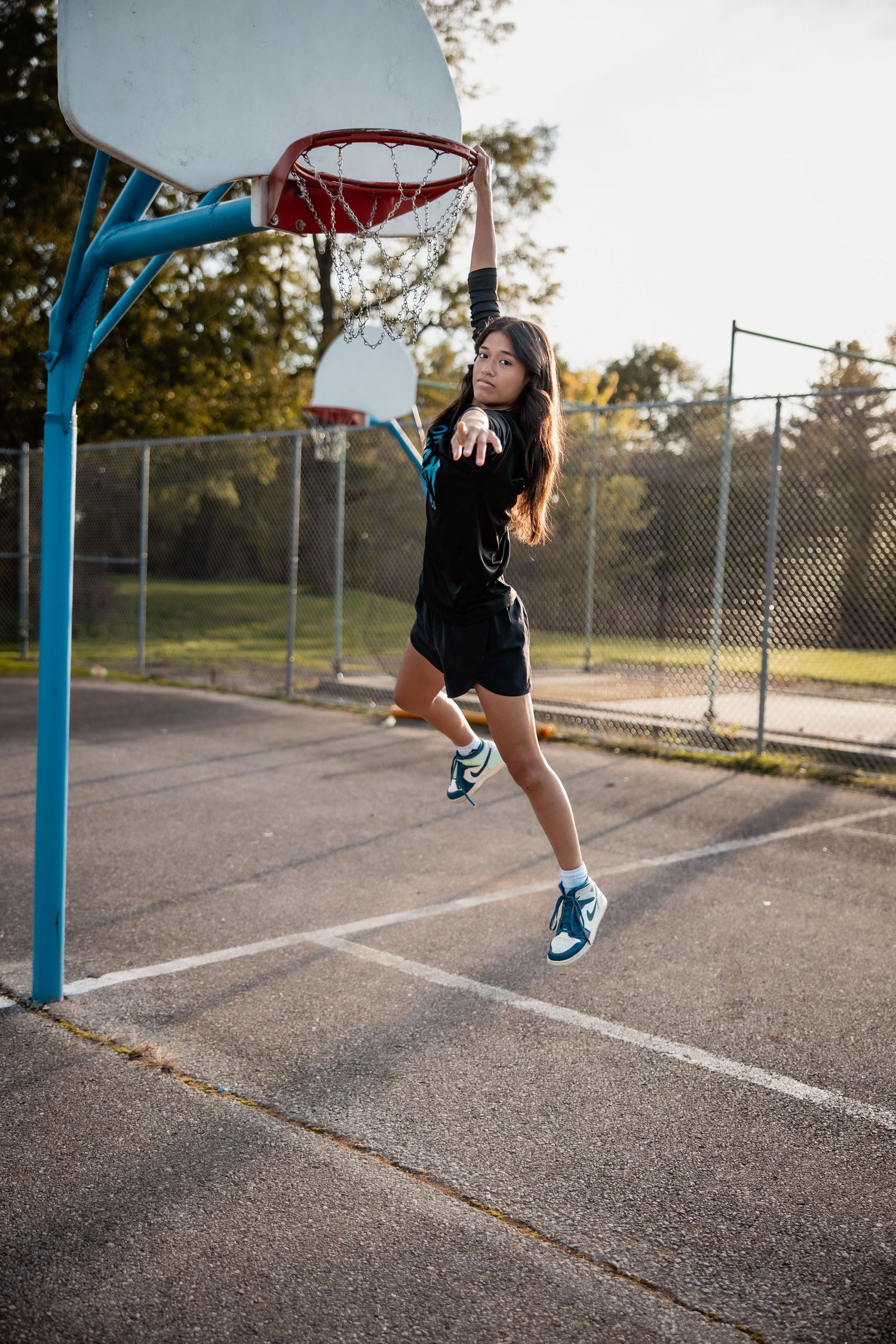 Young woman dunking a basketball on an outdoor court, wearing black shorts and teal sneakers on a sunny day.