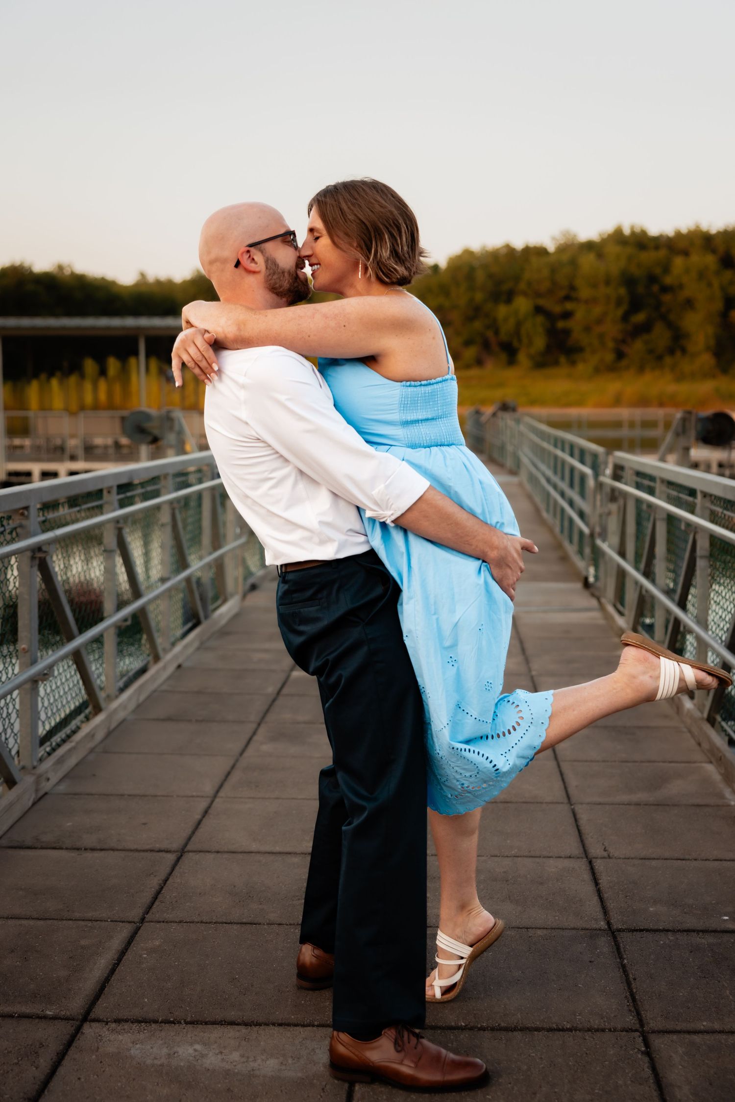 Couple sharing a romantic kiss on a bridge at sunset, man lifting woman in blue dress, lush green trees in background.