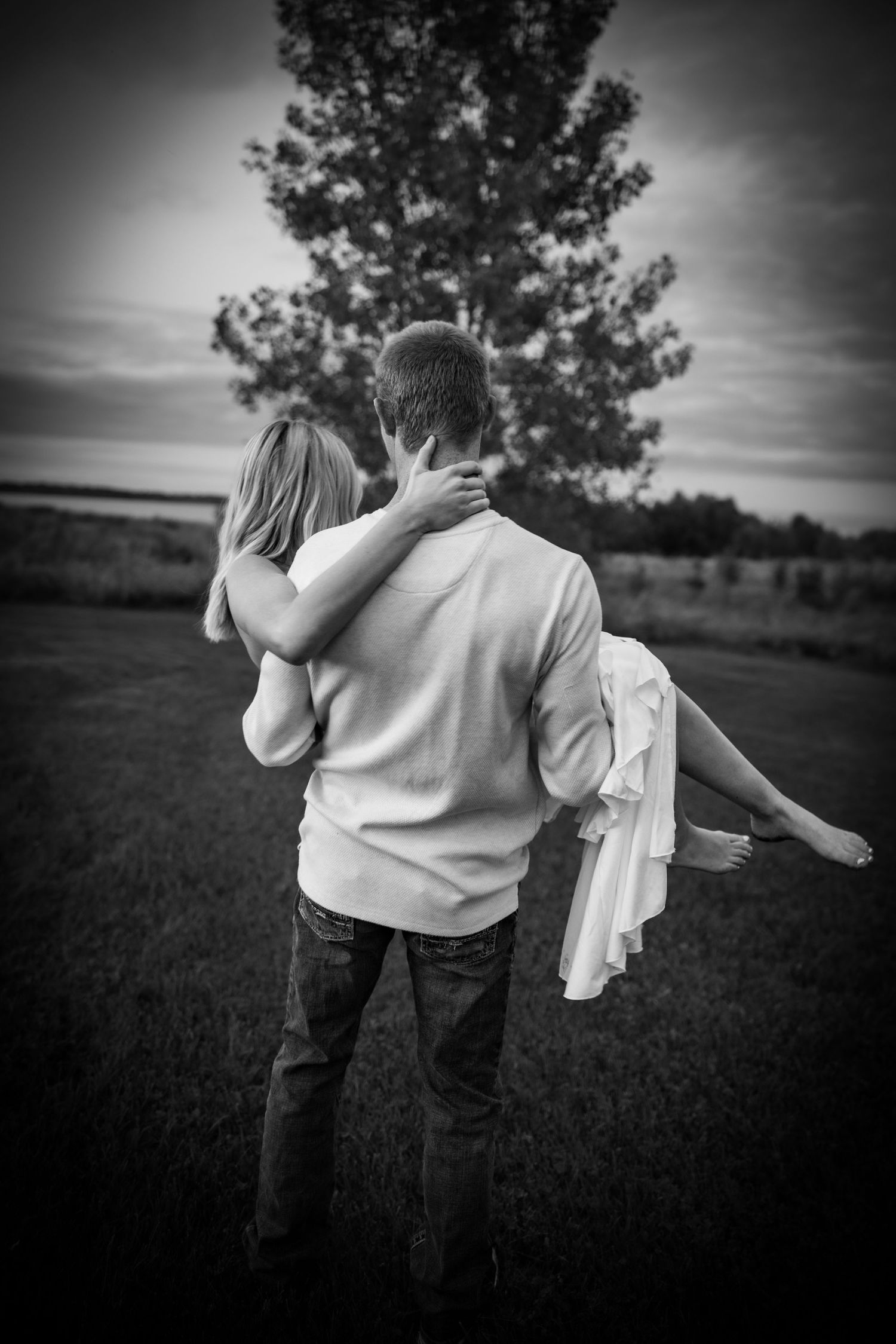 Black and white photo of a man carrying a woman in his arms in a field with a large tree in the background.