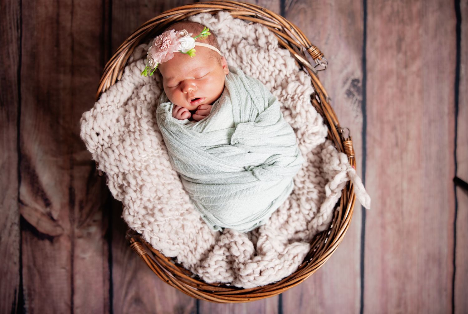 Newborn baby swaddled in mint wrap, sleeping in a wicker basket with a floral headband on rustic wood.