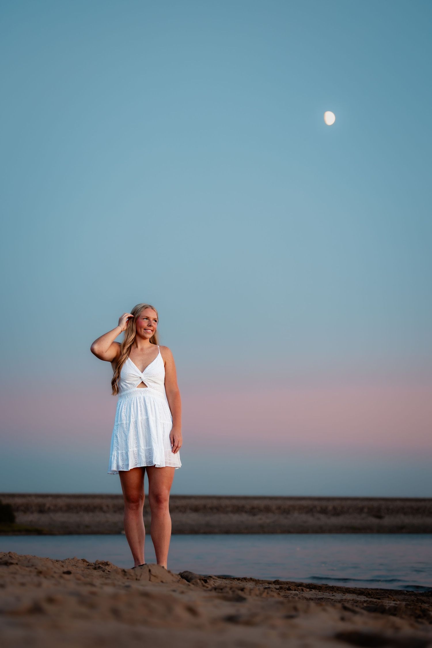 Woman in white dress standing barefoot on beach at twilight with moon visible in blue-pink sky.