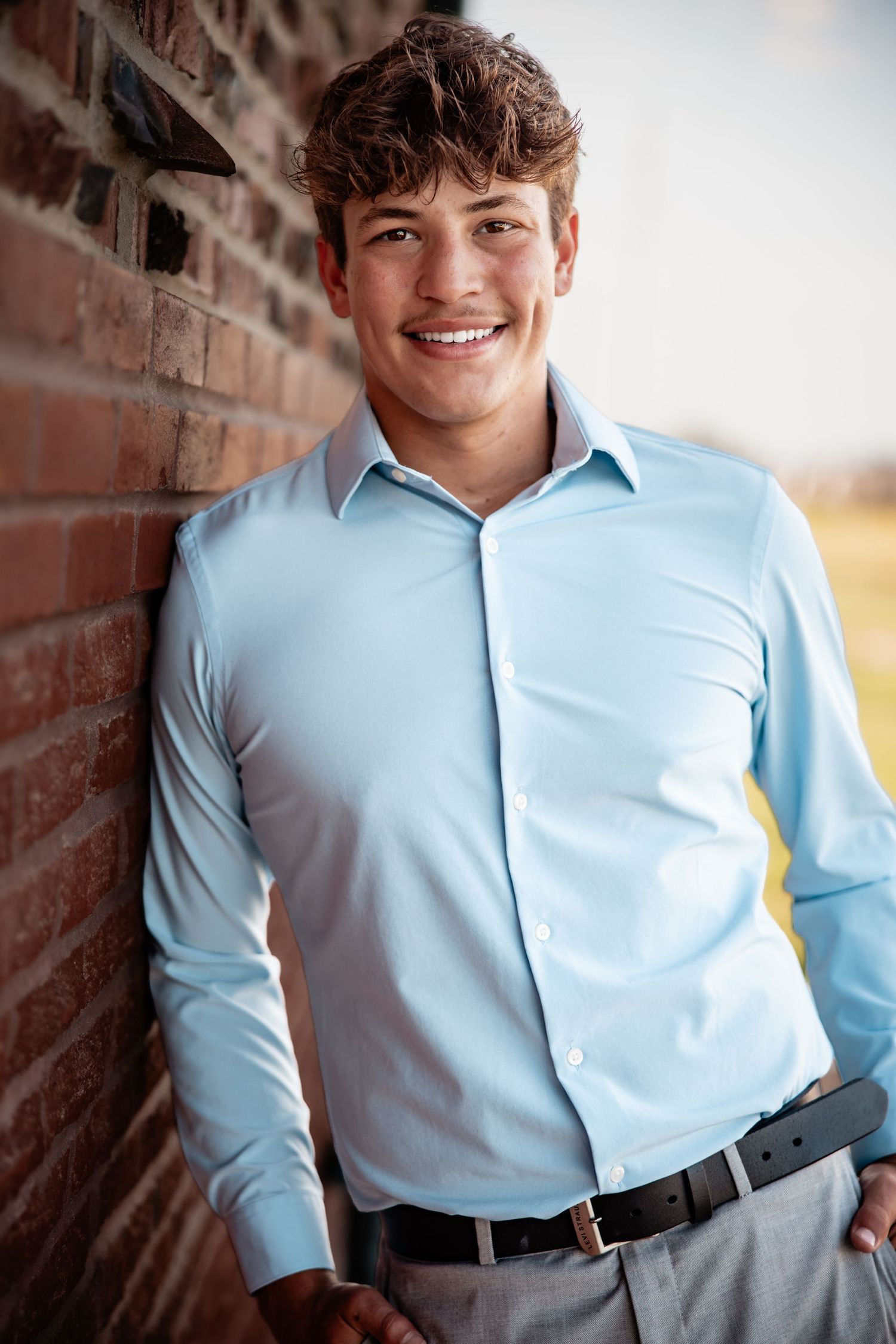 Smiling young man in light blue dress shirt leaning against brick wall outdoors.