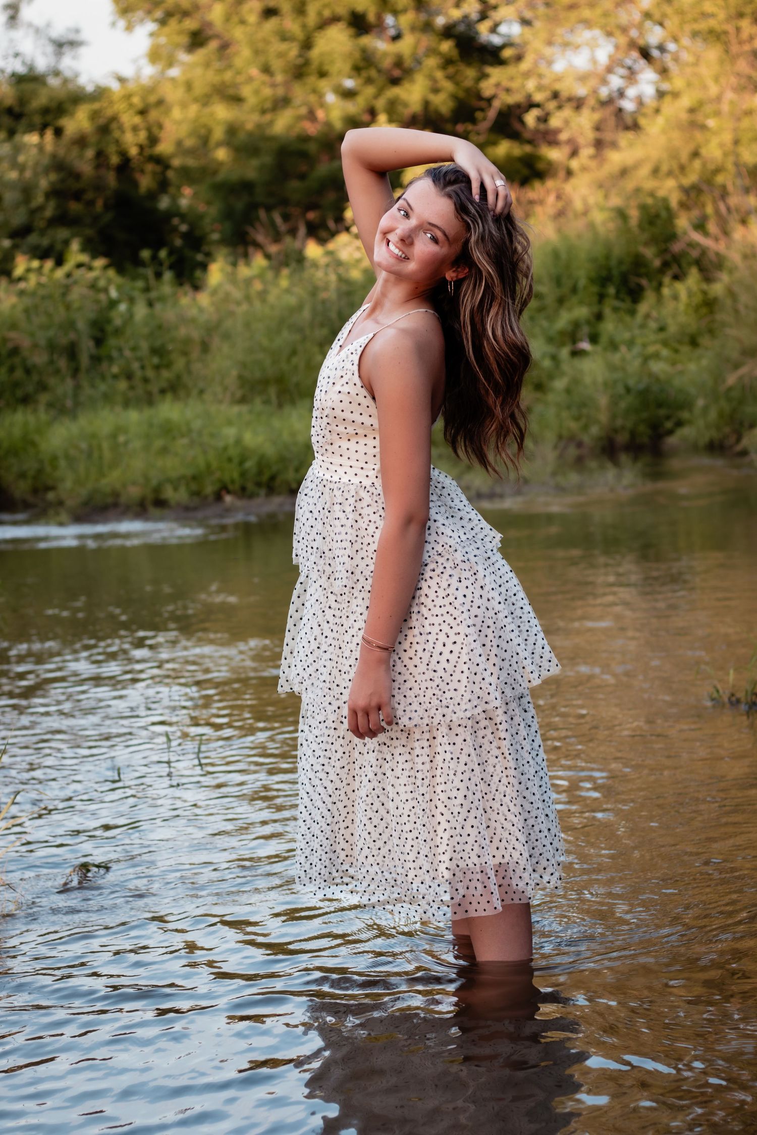 Young woman in white polka dot dress standing in shallow creek, smiling with hand in hair, lush green trees behind her.