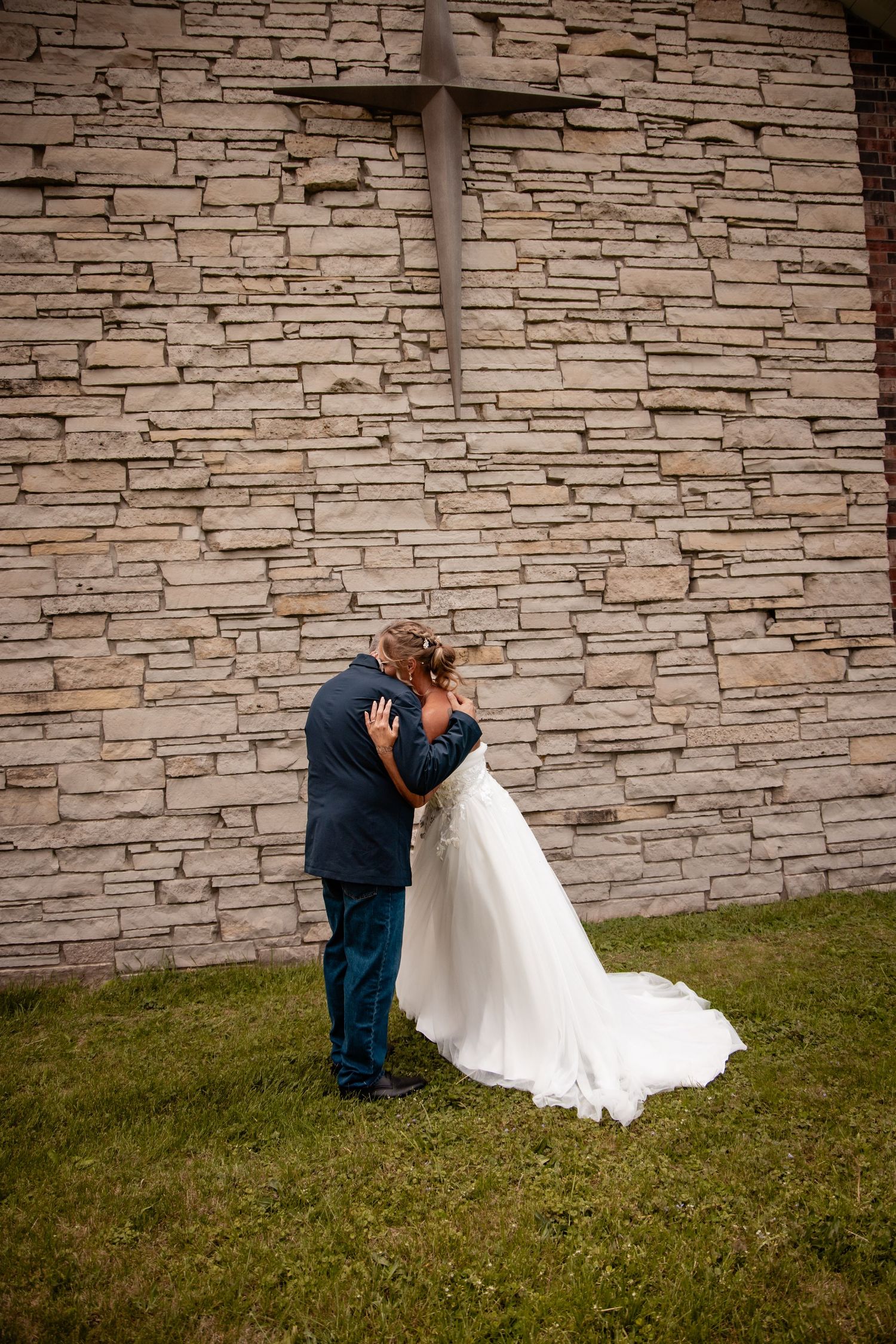 Bride and groom sharing first look embrace against stone church wall with cross.
