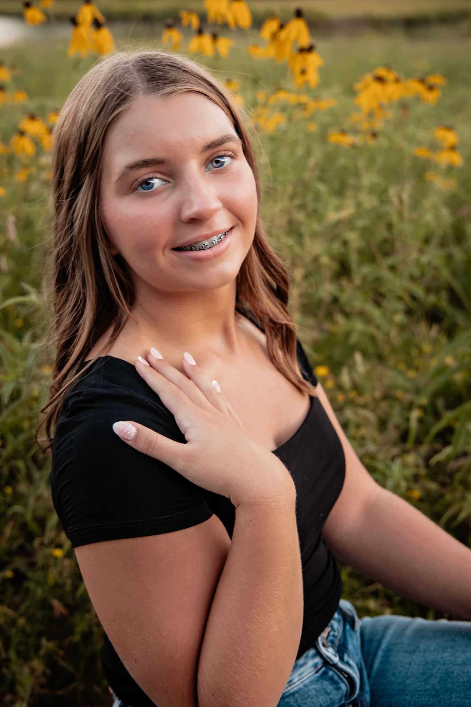 Teen girl with braces smiling in a sunflower field, wearing a black top and jeans, posing outdoors.