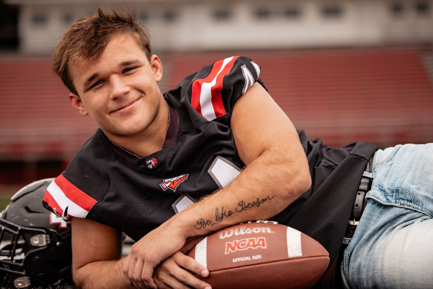 A young male football player in a black and red jersey poses with a Wilson NCAA football on a stadium field.