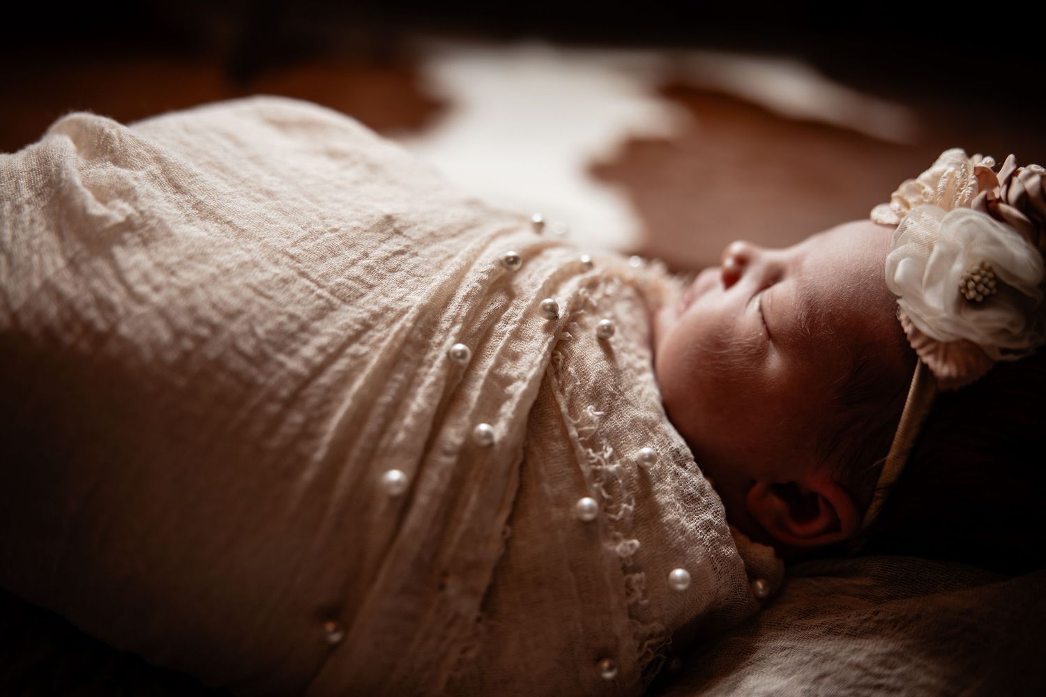 Newborn baby swaddled in a lace wrap with pearl details, wearing a floral headband, sleeping peacefully.