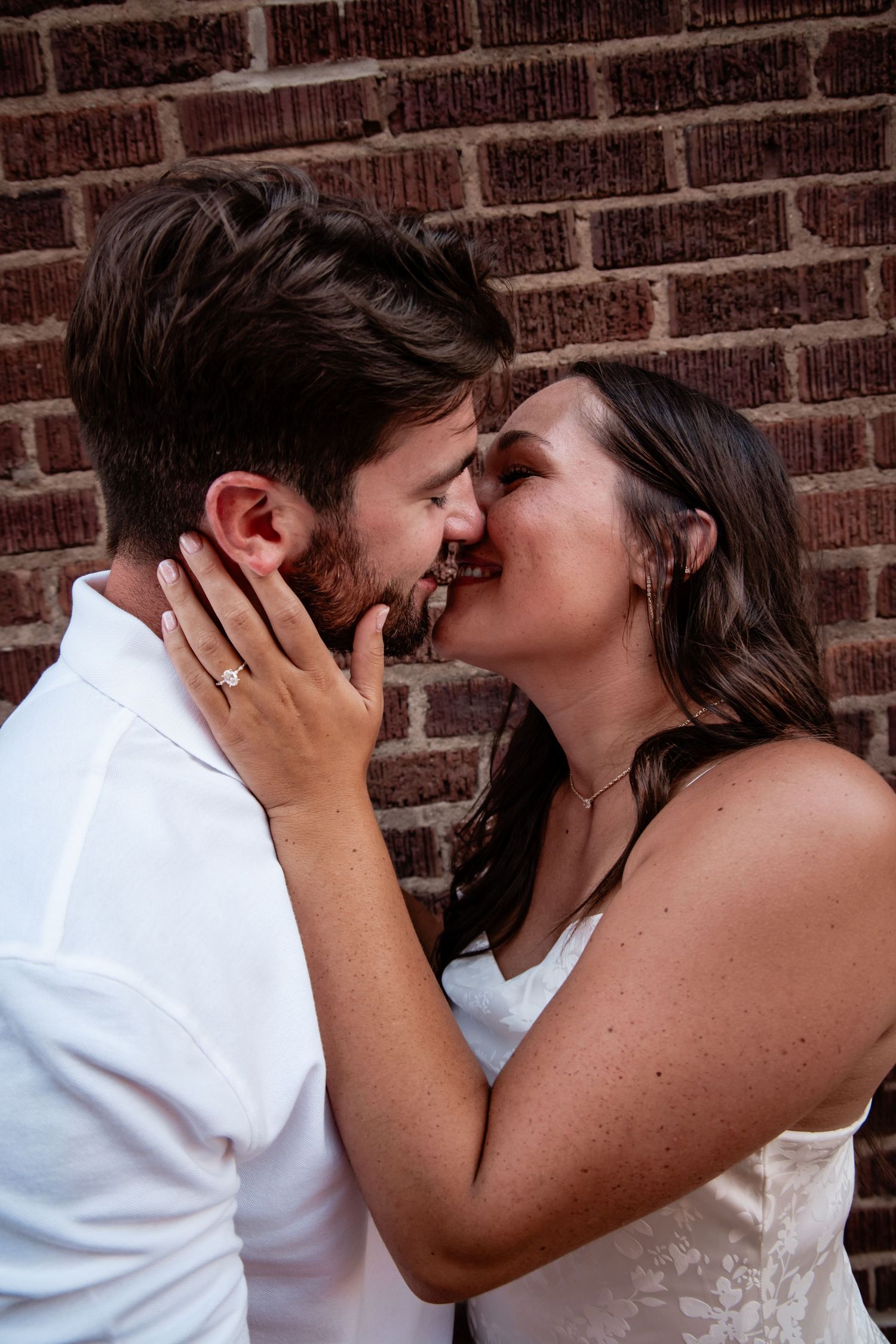 Couple sharing a romantic kiss against a brick wall, woman wearing an engagement ring in a tender embrace.