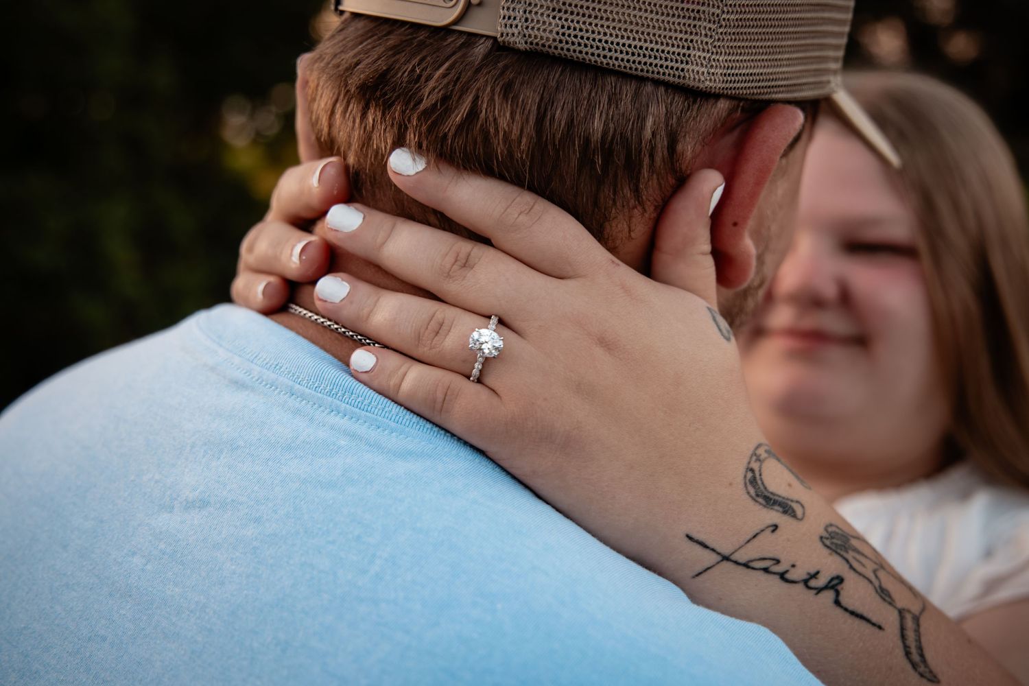 Woman showing off engagement ring with faith tattoo on wrist while embracing partner outdoors.