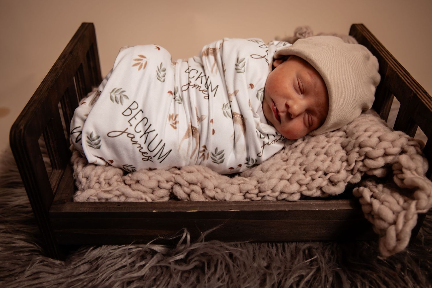 Newborn baby swaddled in white Blokum blanket sleeping in rustic wooden mini crib with chunky knit blanket.