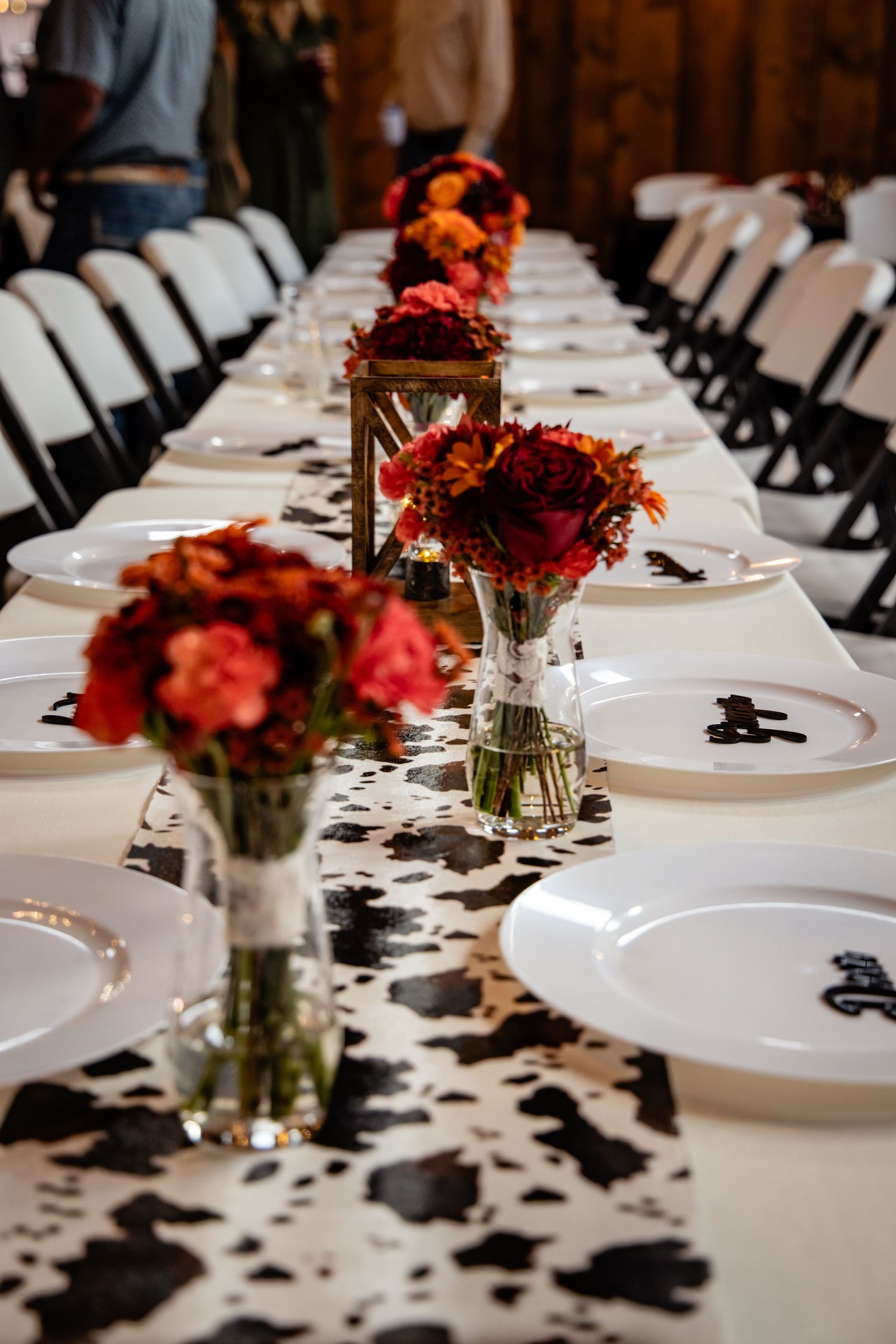 Rustic wedding reception table with cow print runner, red and orange floral centerpieces, white plates, and candle lanterns.