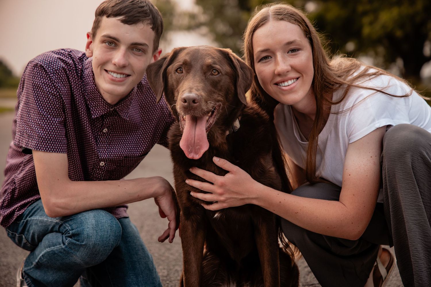 Young couple smiling and posing with a chocolate Labrador dog outdoors in a warm, softly lit setting.