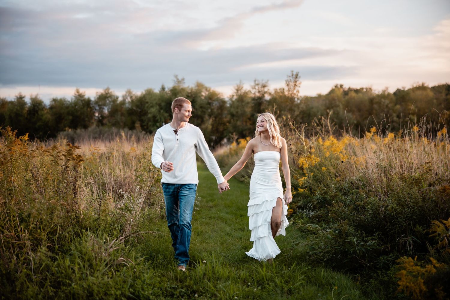 A newlywed couple holds hands walking through a lush green meadow at golden hour in their wedding attire.