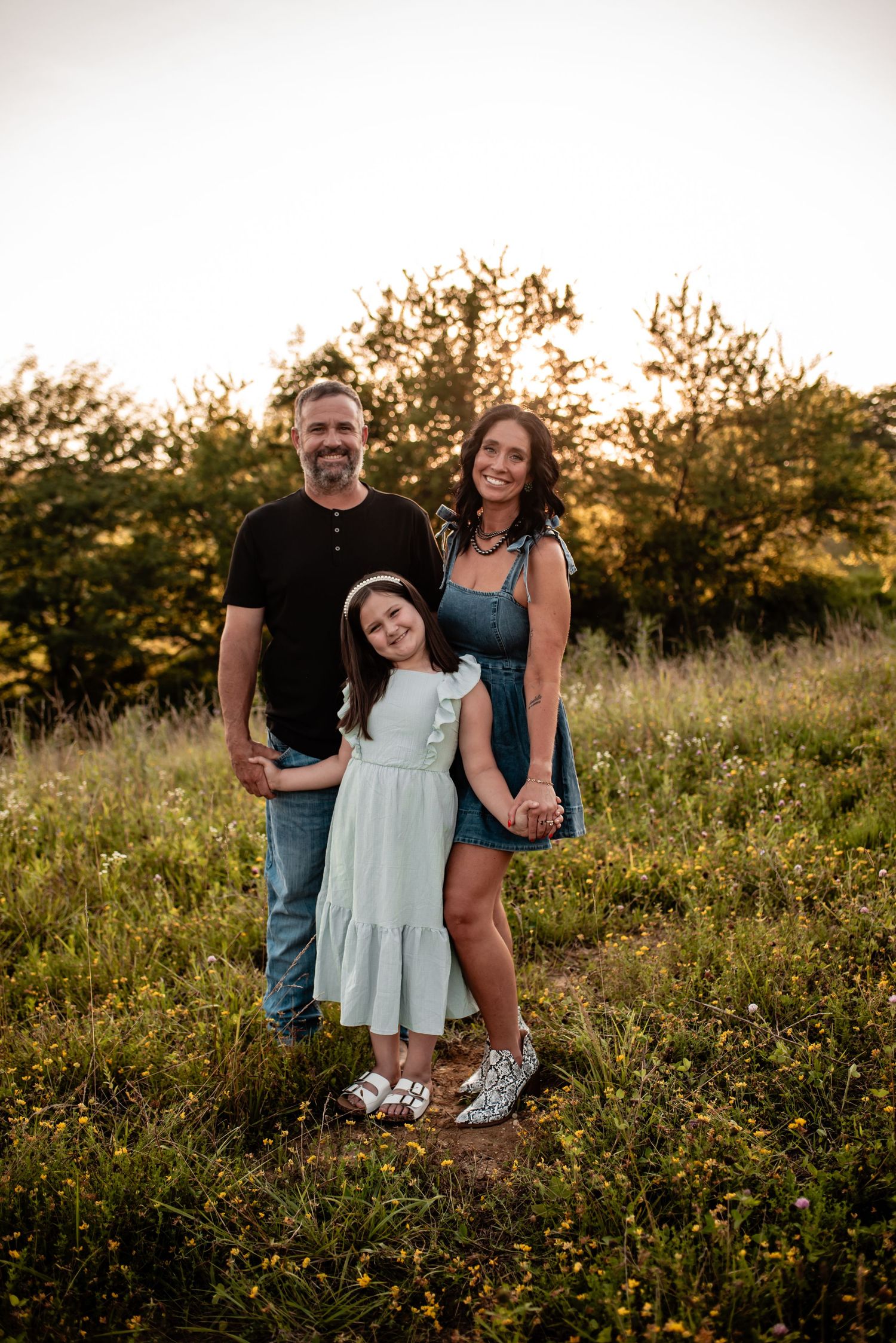 A smiling family of three poses in a sunlit meadow, with parents flanking their young daughter in a light green dress.