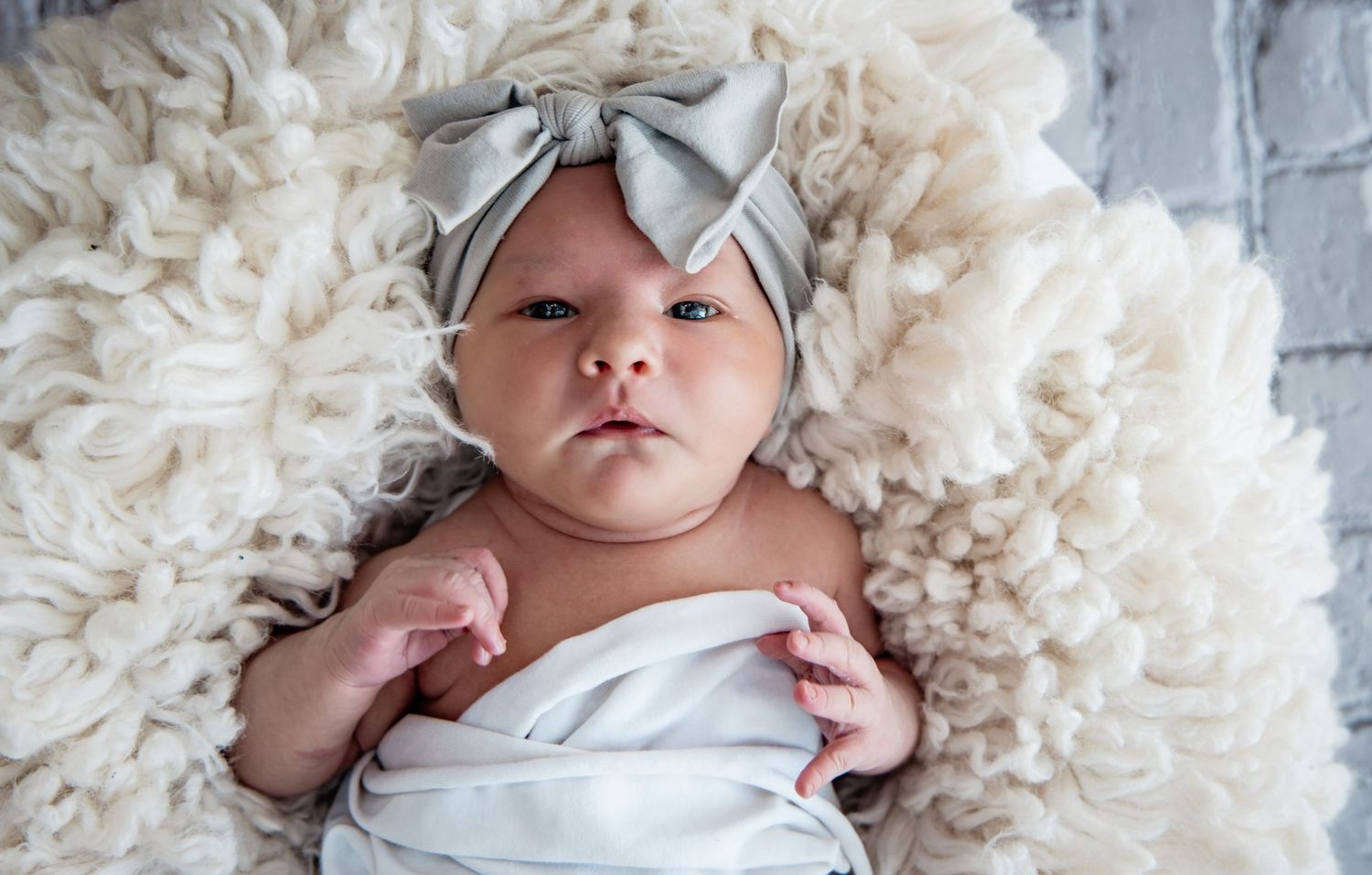 Newborn baby wrapped in white swaddle with gray bow headband, resting on fluffy cream wool blanket.