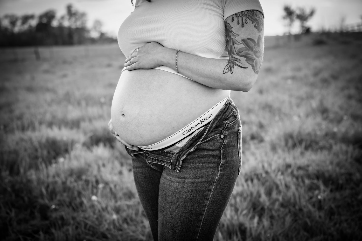 Black and white maternity photo of pregnant woman cradling baby bump in a grassy field.