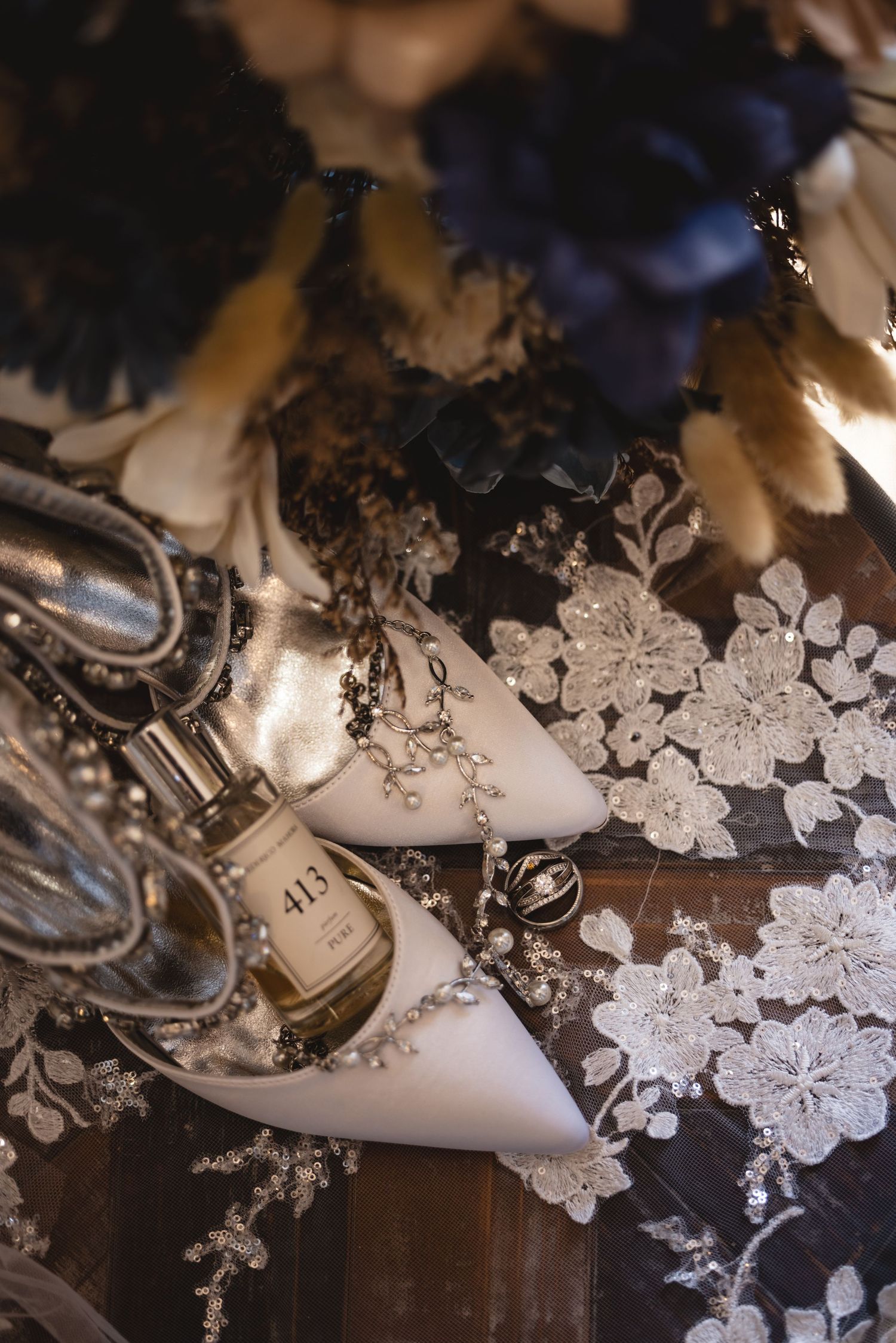 Bridal flat lay featuring white pearl-embellished heels, perfume bottle, rings, and dark floral bouquet.