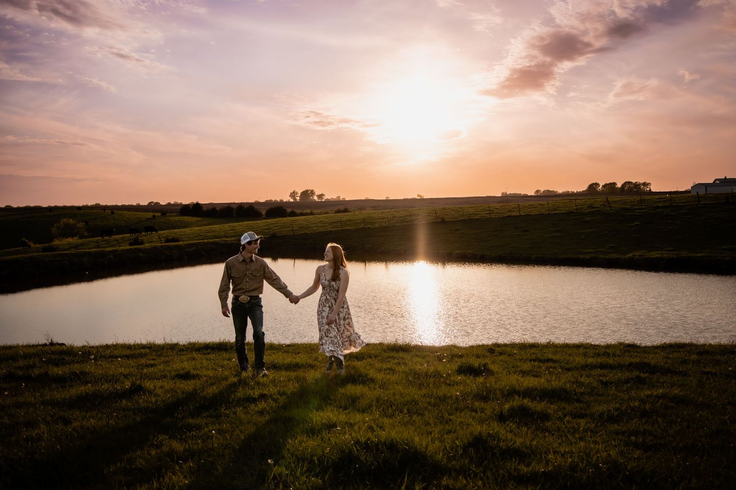 Couple holding hands at sunset by a serene pond in a lush green field.