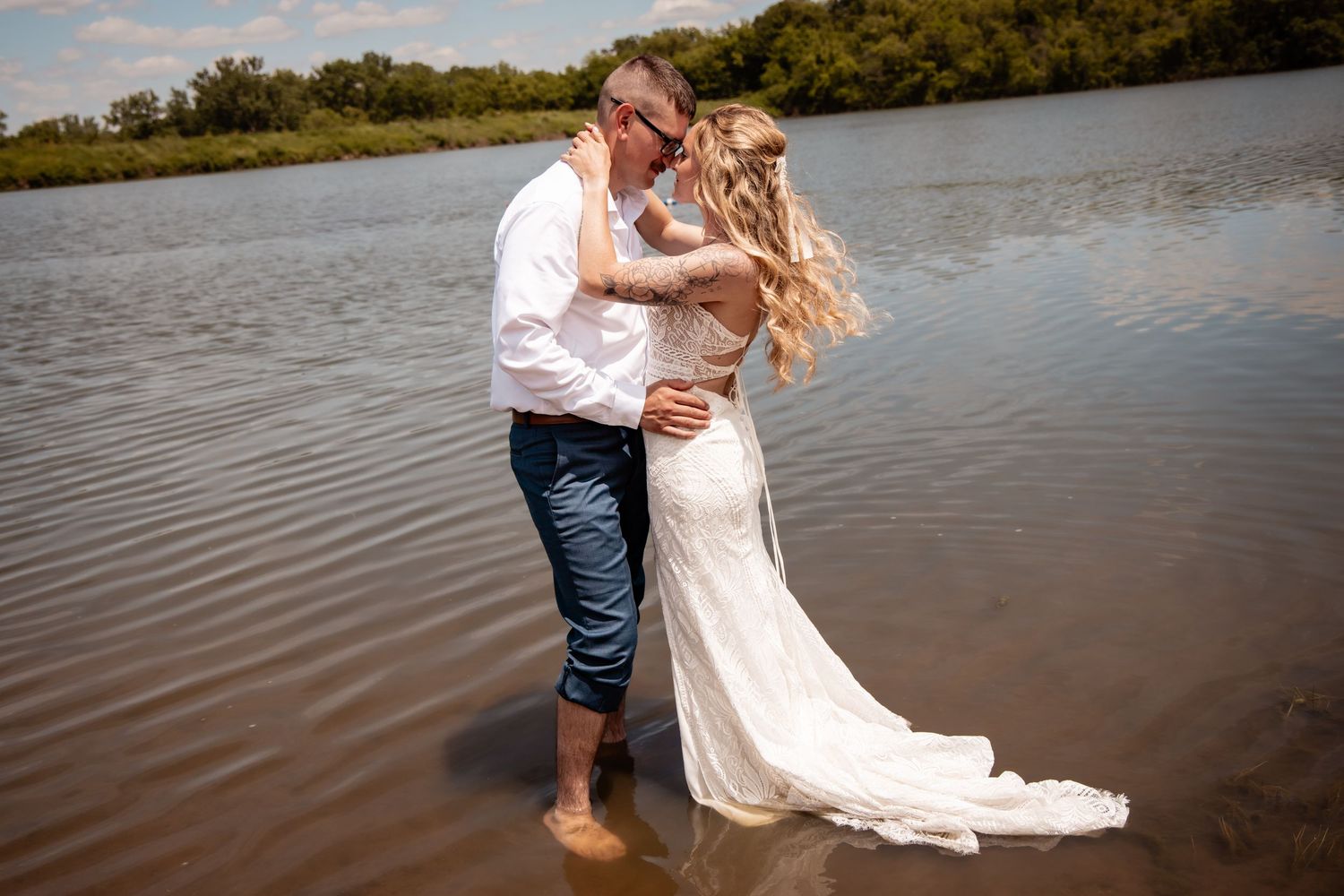 Bride and groom standing in shallow water, sharing a romantic embrace in wedding attire near a serene lake.