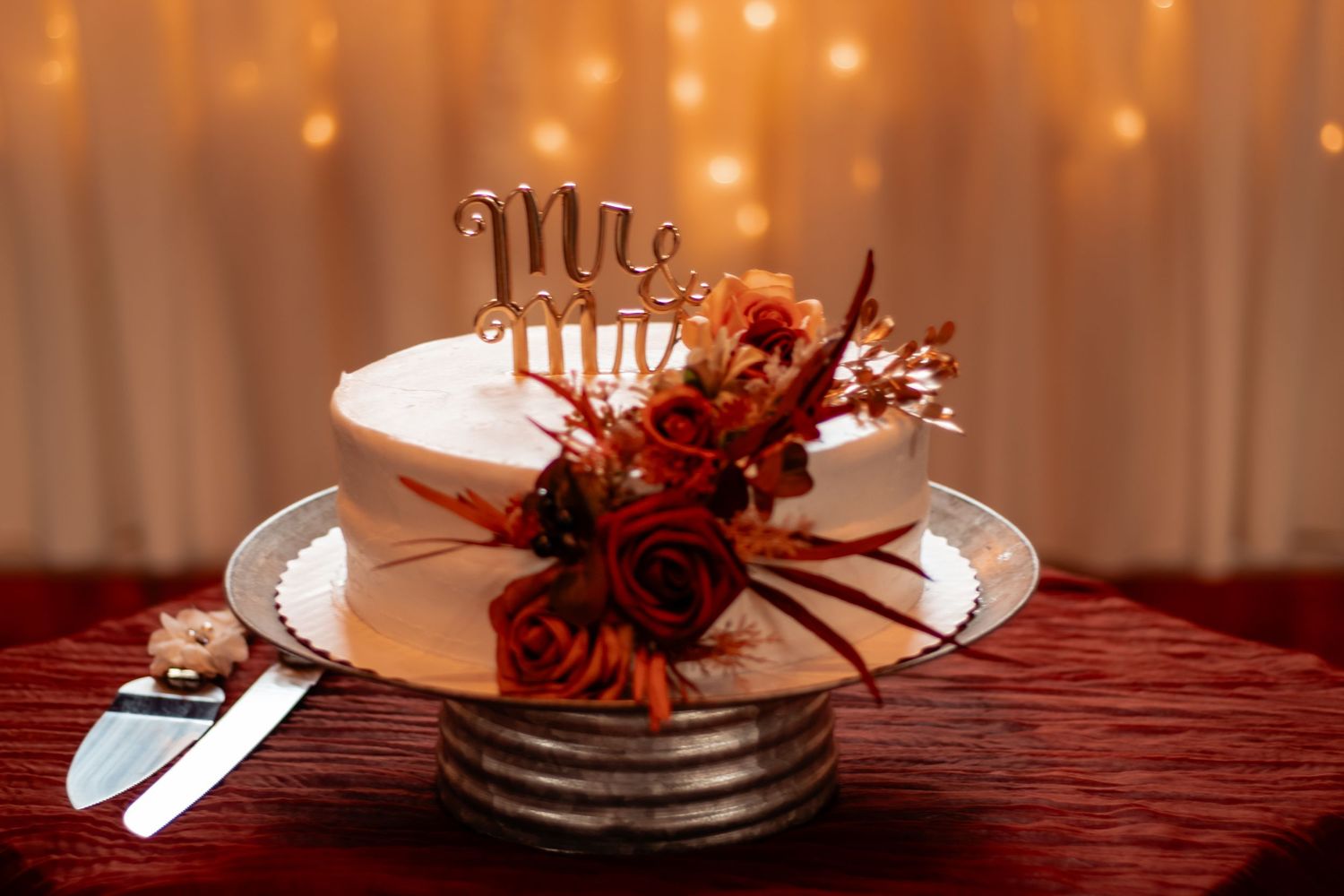 White wedding cake with red floral decor and Mr. & Mrs. topper on silver stand against warm bokeh backdrop.