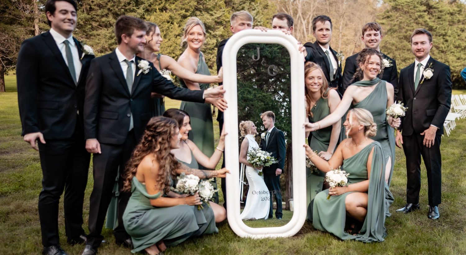Wedding party poses outdoors holding a mirror reflecting the bride and groom in a romantic group photo.