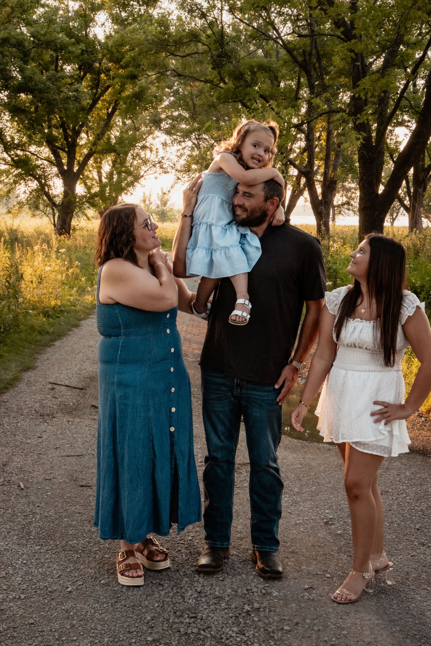 Family of four posing on a tree-lined path at golden hour, with dad holding young girl on shoulders.