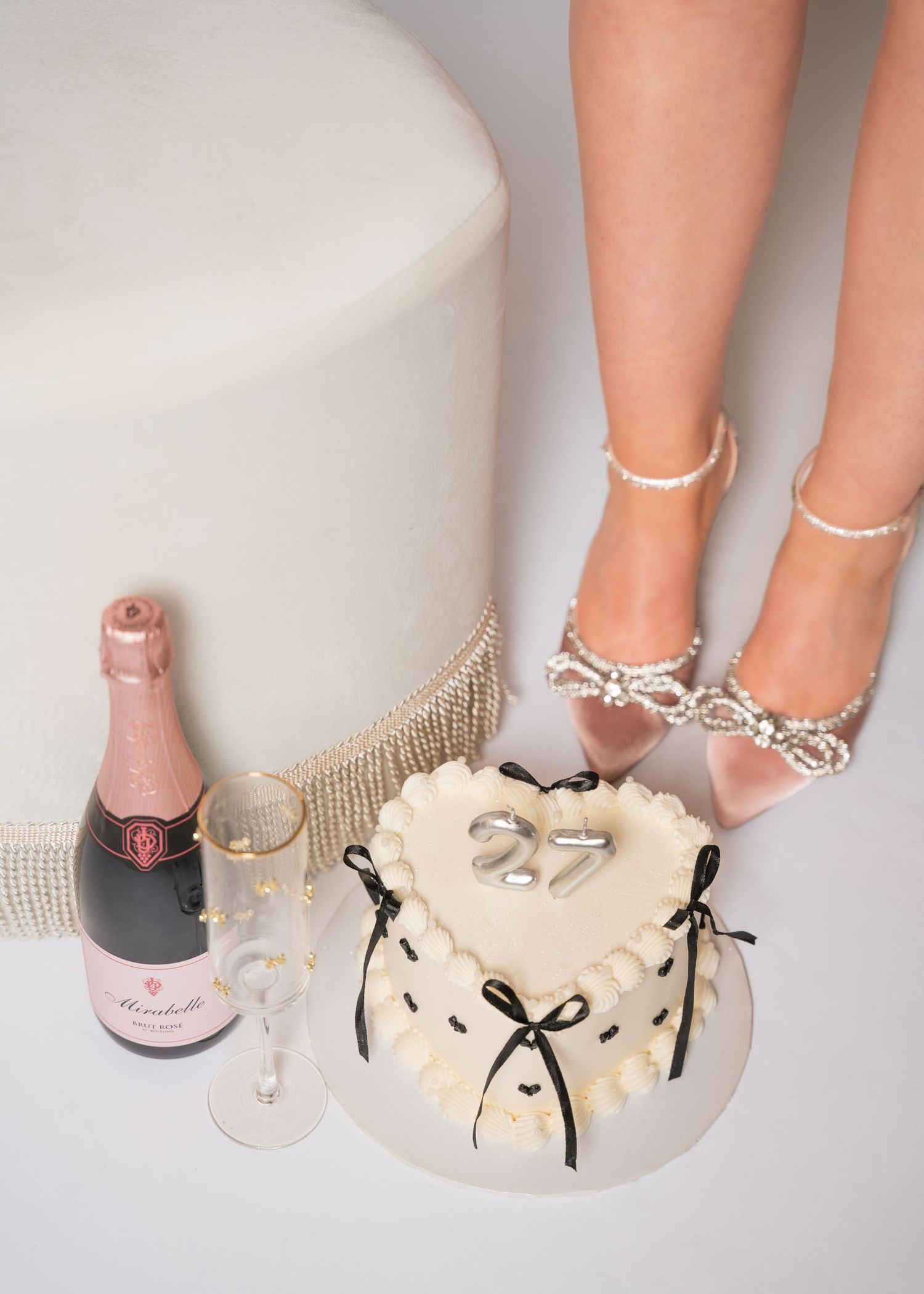 Heart-shaped 27th birthday cake with champagne bottle and pearl heels in a black and white flat lay.