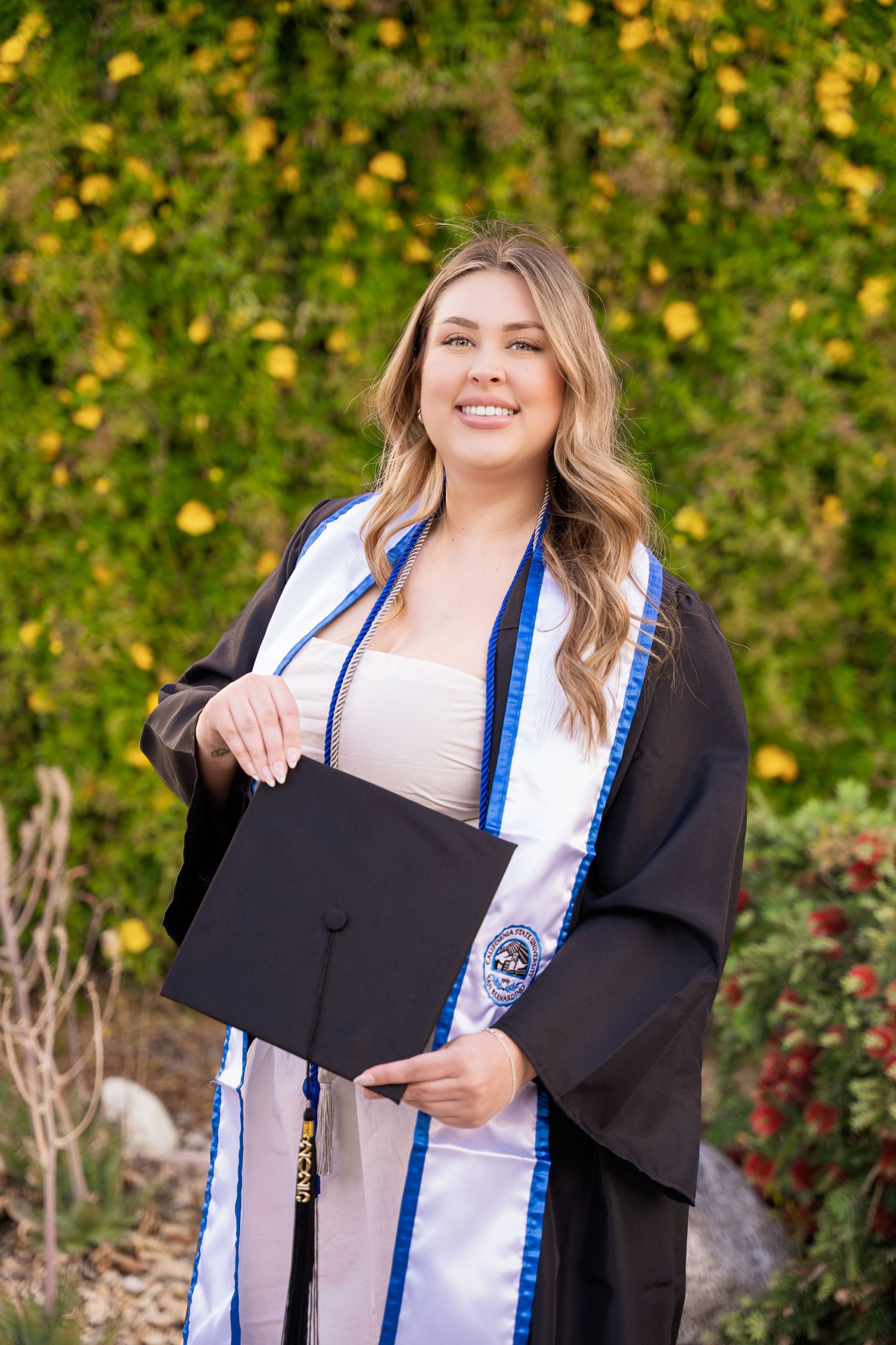 Smiling female graduate in black gown and blue stole holding cap in front of colorful foliage.