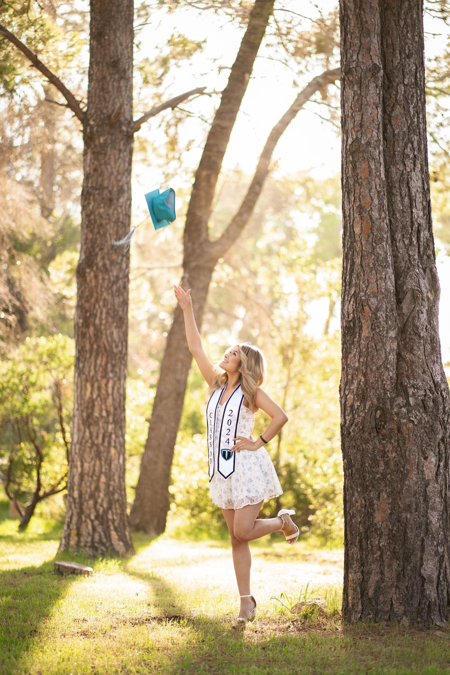 Blonde woman in white dress tossing teal graduation cap among sunlit trees in forest setting.