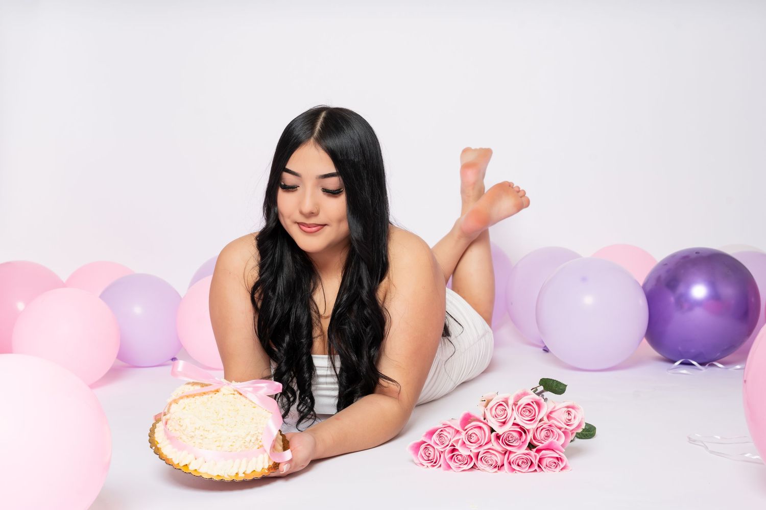Young woman lying on floor with birthday cake and pink roses surrounded by pink and purple balloons.
