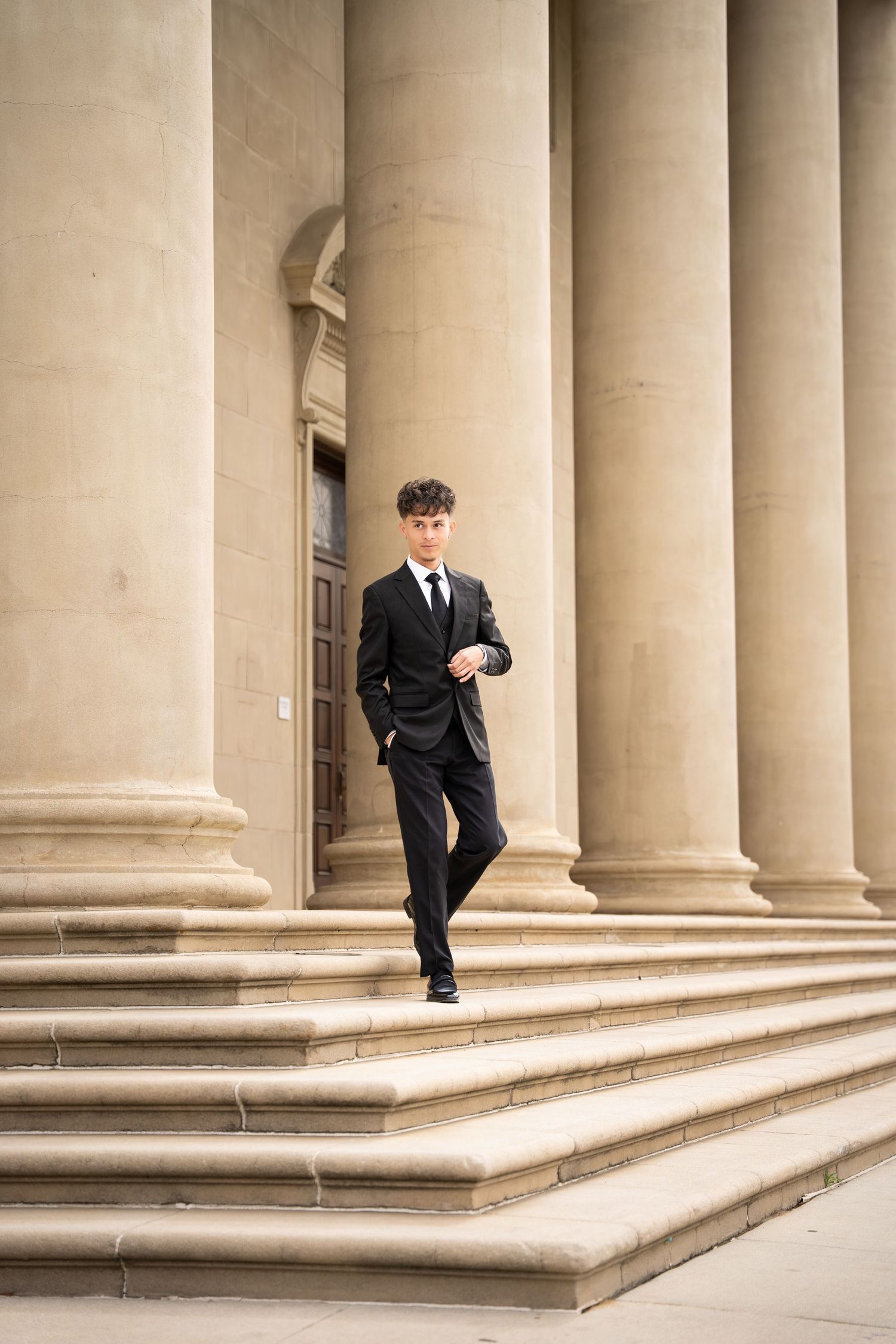 Young man in black suit walks down steps of classical building with tall columns.