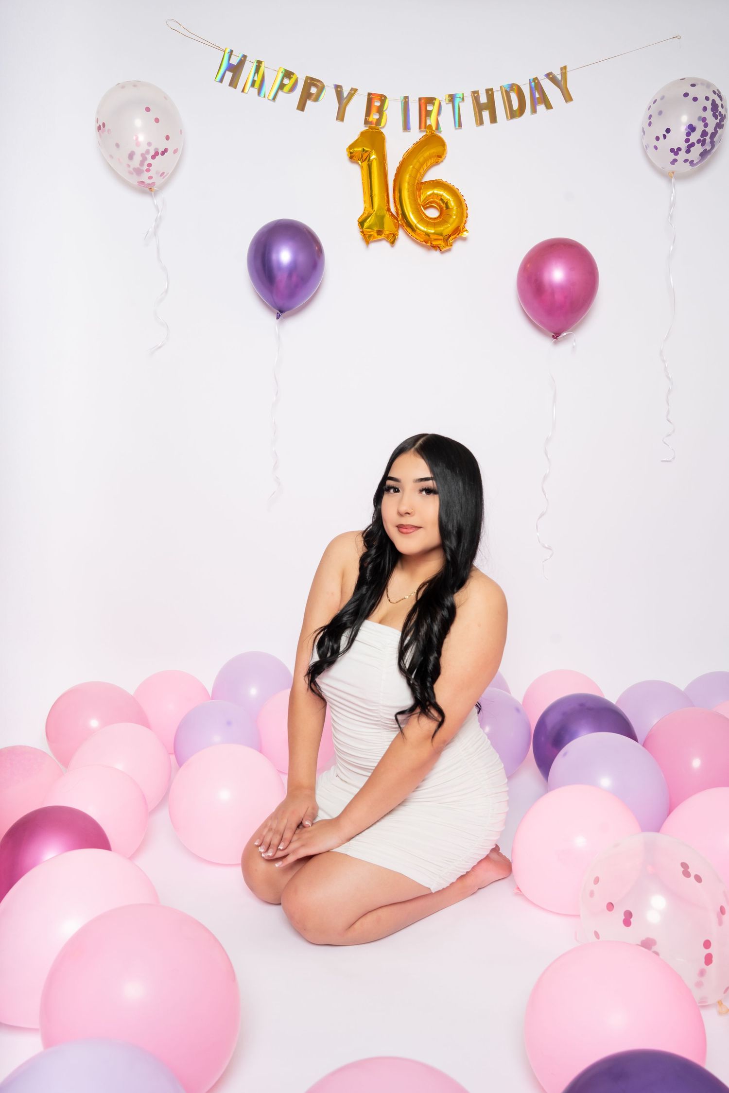Young woman in white dress sitting on floor with Happy Birthday 16 balloons and pink purple balloons.