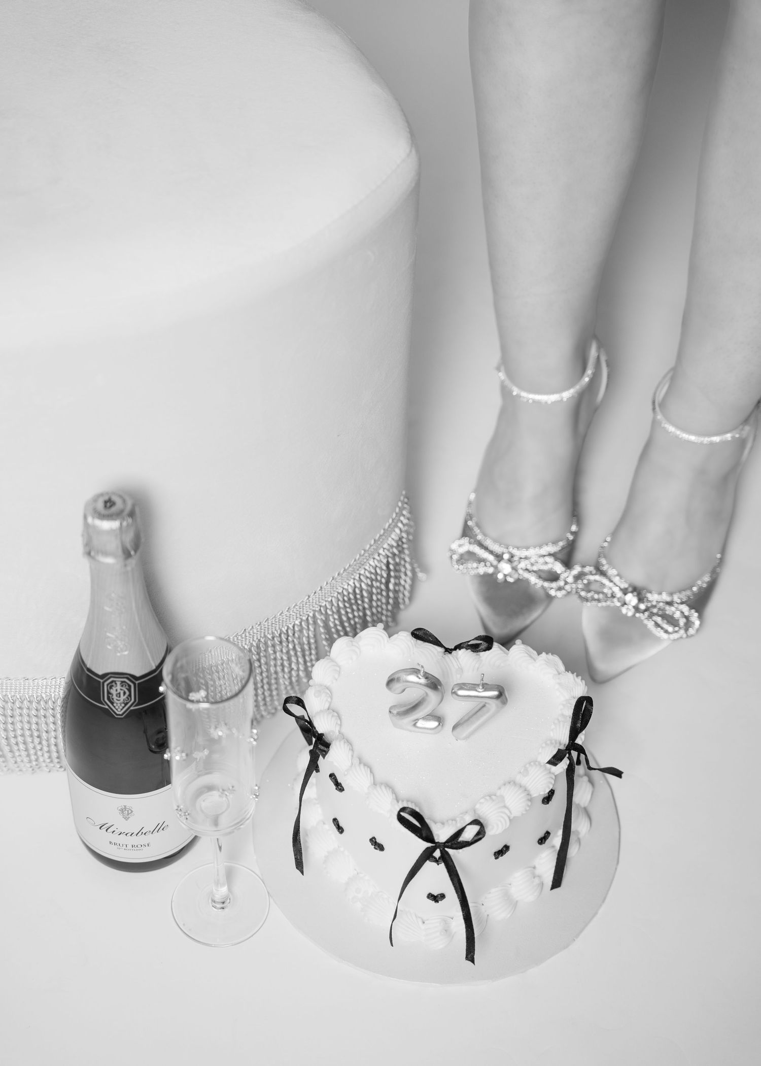Black and white flat lay of heart-shaped birthday cake, champagne, and pearl heels on studio floor.