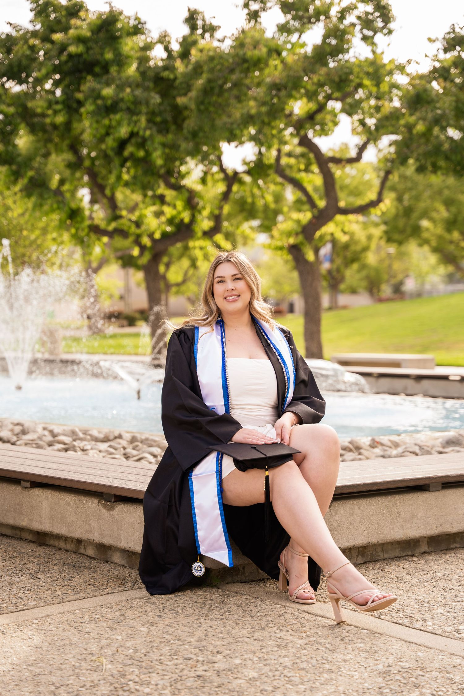 Female graduate in black gown and blue stole sits by fountain for CSUSB graduation photos.