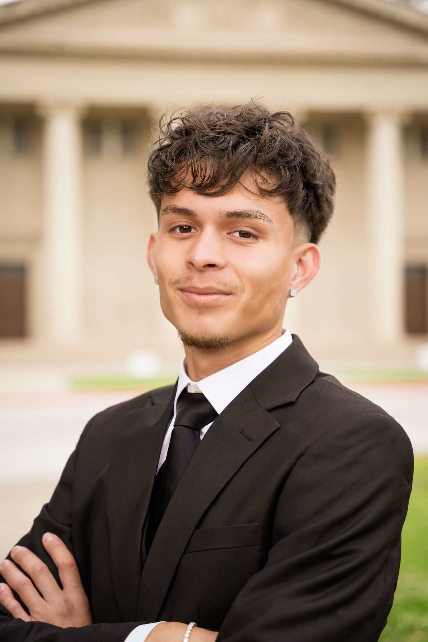 Smiling young man in black suit and tie poses with arms crossed outside university.