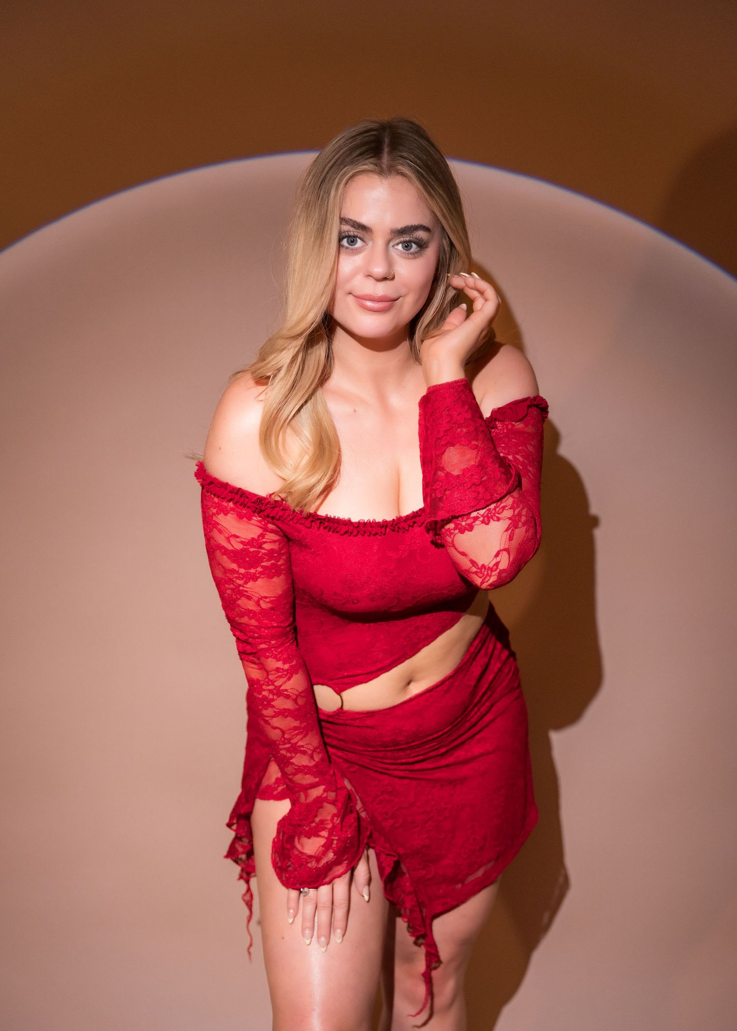 Blonde woman in red lace two-piece outfit with off-shoulder top posing in a warmly lit studio.