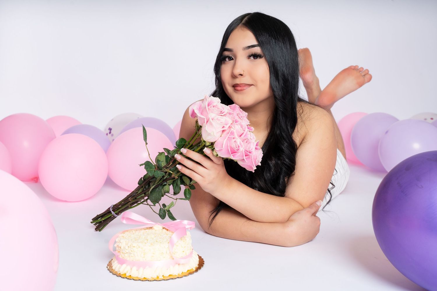 Young woman lying down holding pink roses bouquet with birthday cake and colorful balloons on white background.