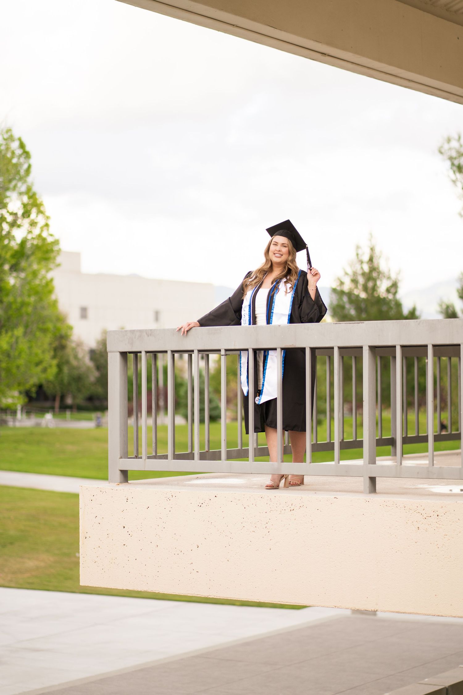 Female graduate in cap and gown posing on an outdoor campus balcony, holding diploma proudly.