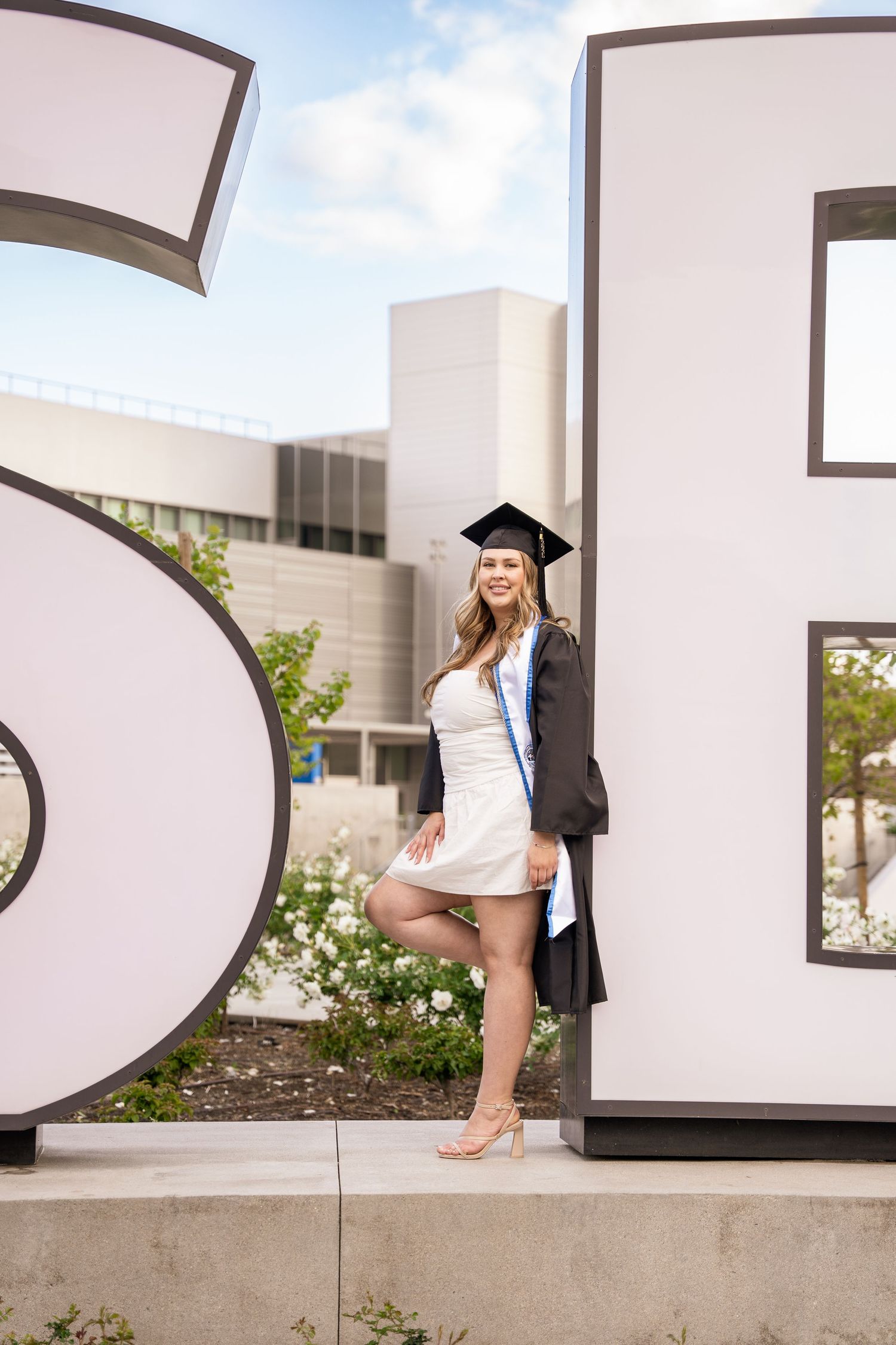 Female graduate in cap and gown posing confidently beside large campus letter signage outdoors.