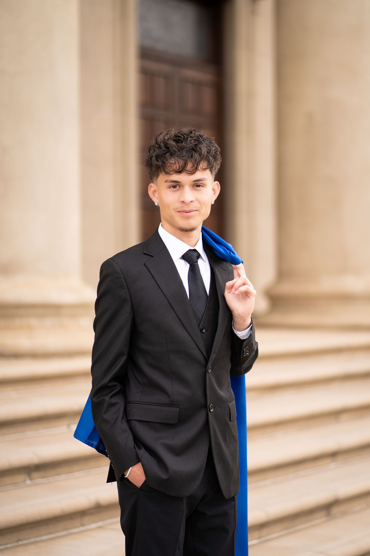 Young male graduate in black suit holds blue graduation stole on university steps.