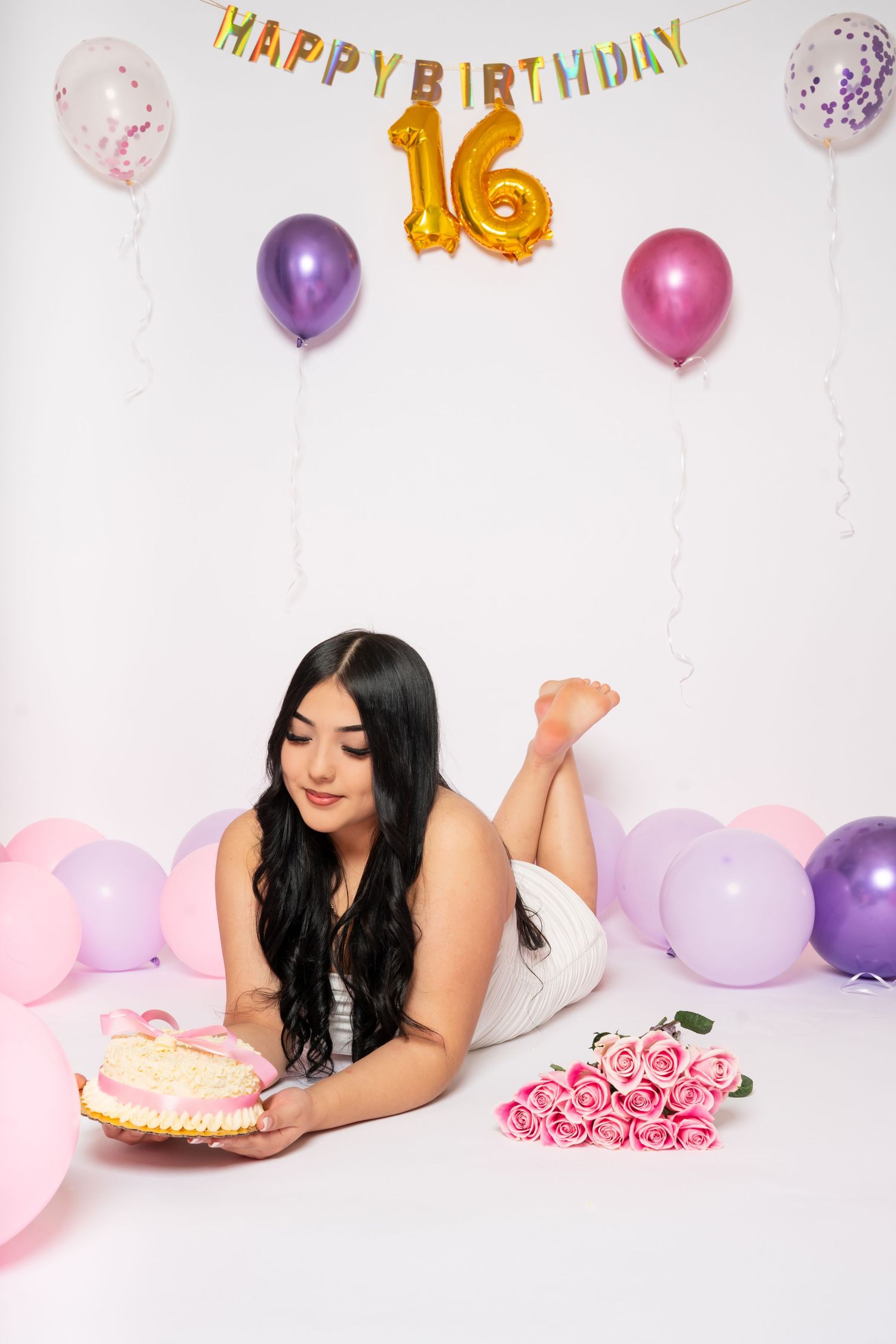 Young woman lying on floor with birthday cake near Happy Birthday 16 sign and colorful balloons.