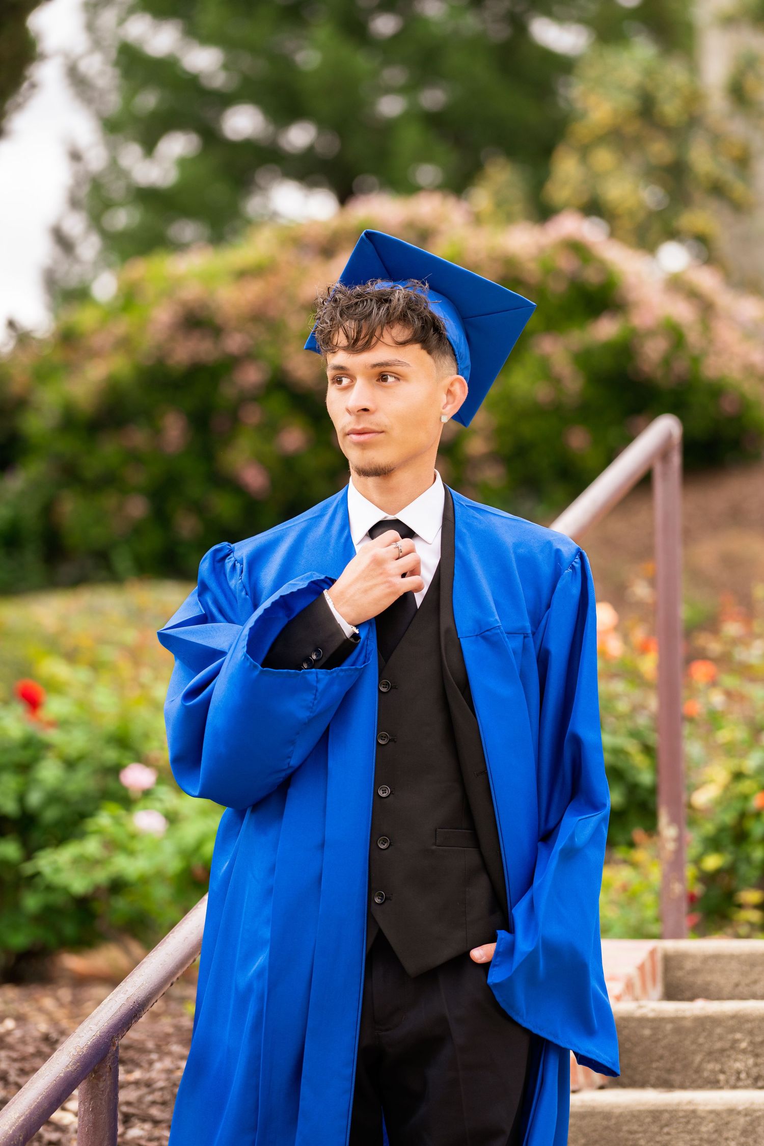 Male graduate in blue cap and gown adjusting tie on outdoor stairway with garden background.