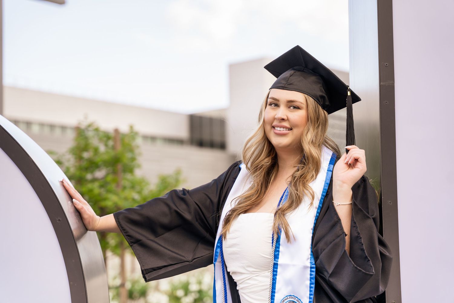 Female graduate in black cap and gown poses by modern campus architecture at CSUSB.