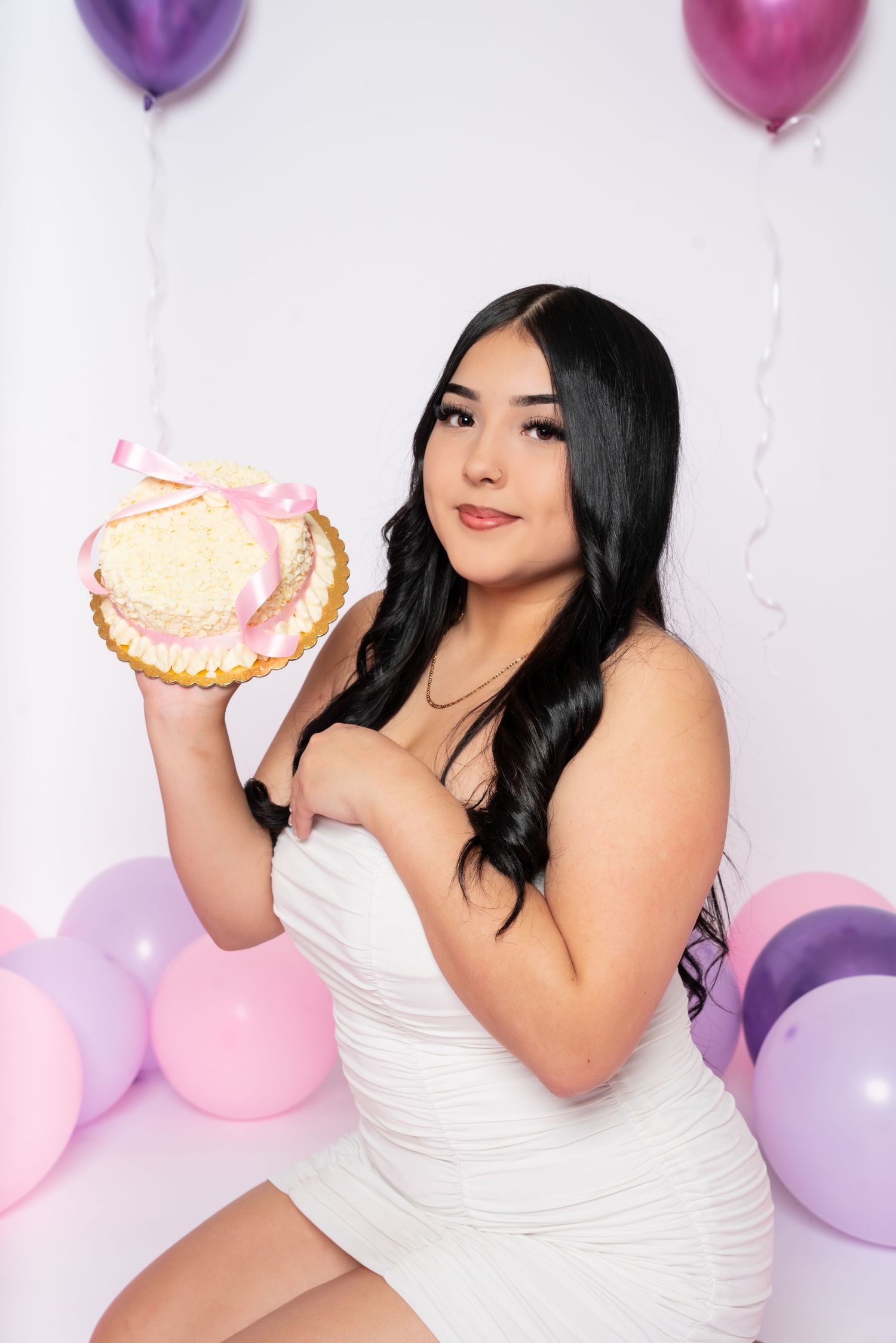 Young woman in white dress sitting holding birthday cake with pink and purple balloons behind her.