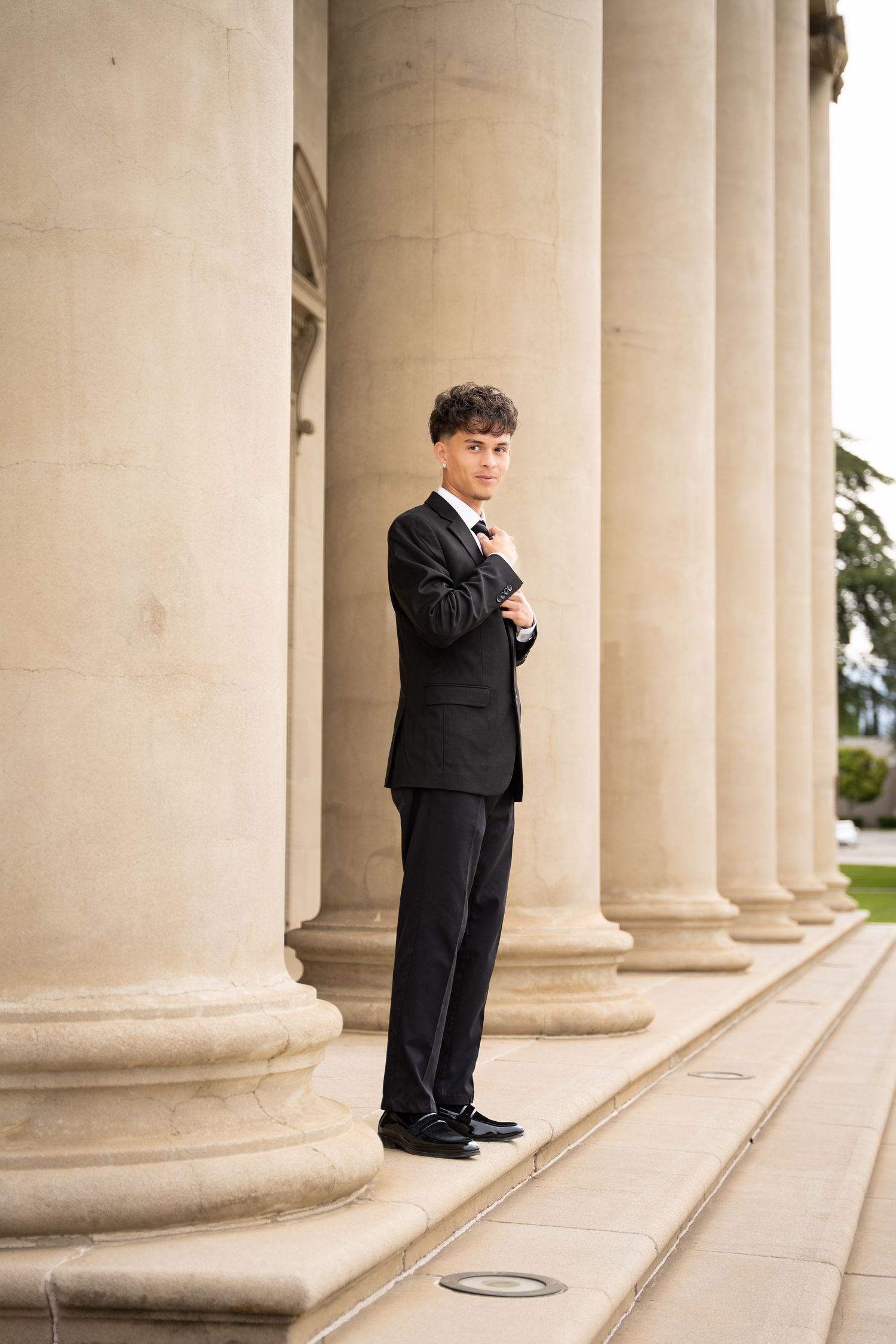 Young man in black suit standing with arms crossed beside tall stone columns of classic building.