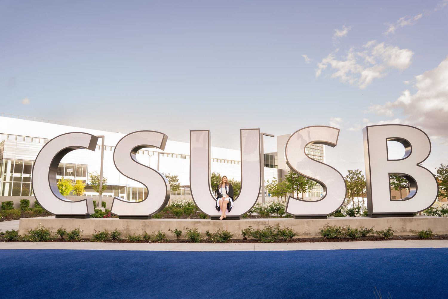 Graduate poses in front of large CSUSB letter sign on blue campus plaza.